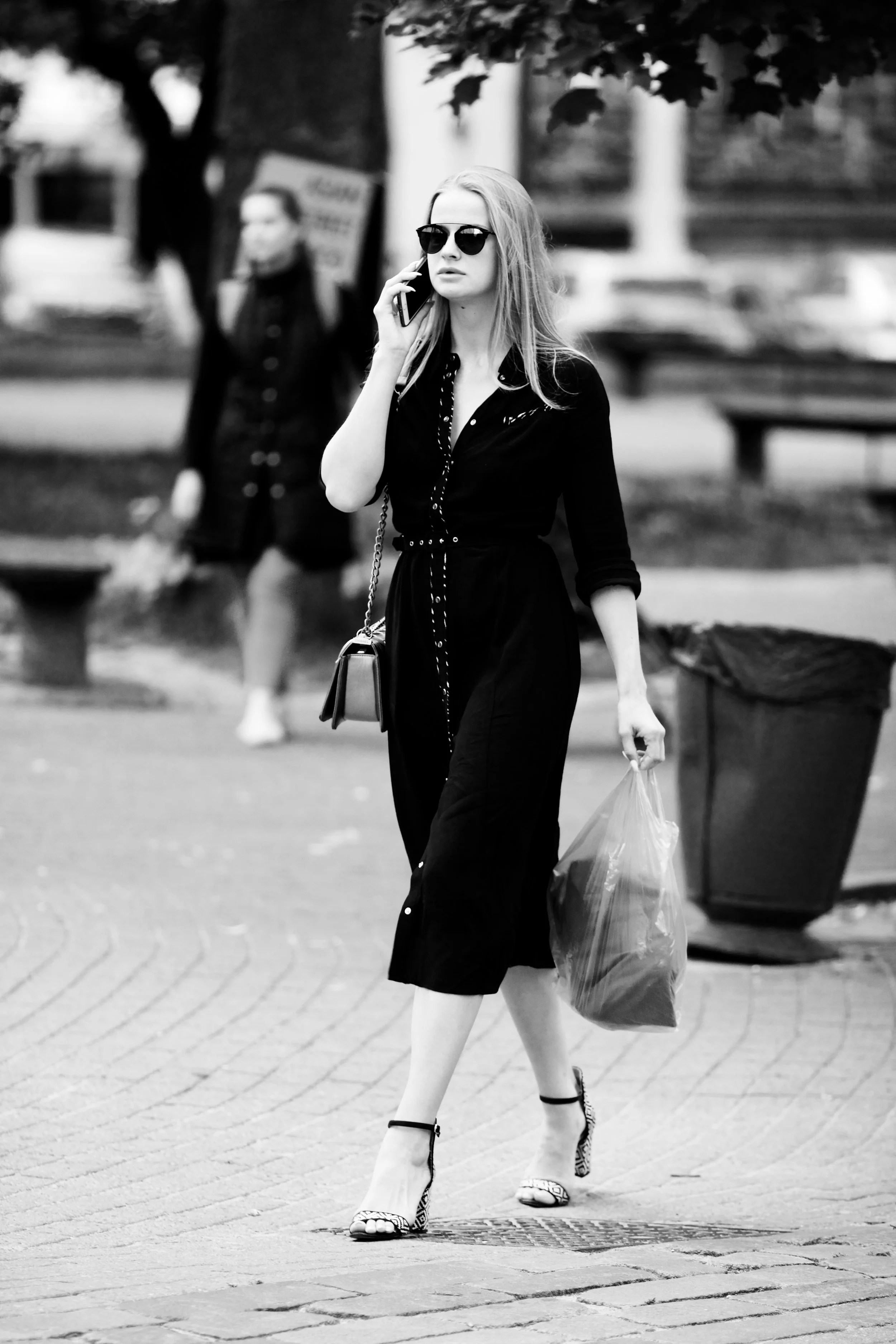 Black and white photo of a woman walking on a sidewalk, talking on her cellphone, wearing sunglasses, a black dress, and high heels, carrying a plastic bag and a purse, with park benches and trees in the background.