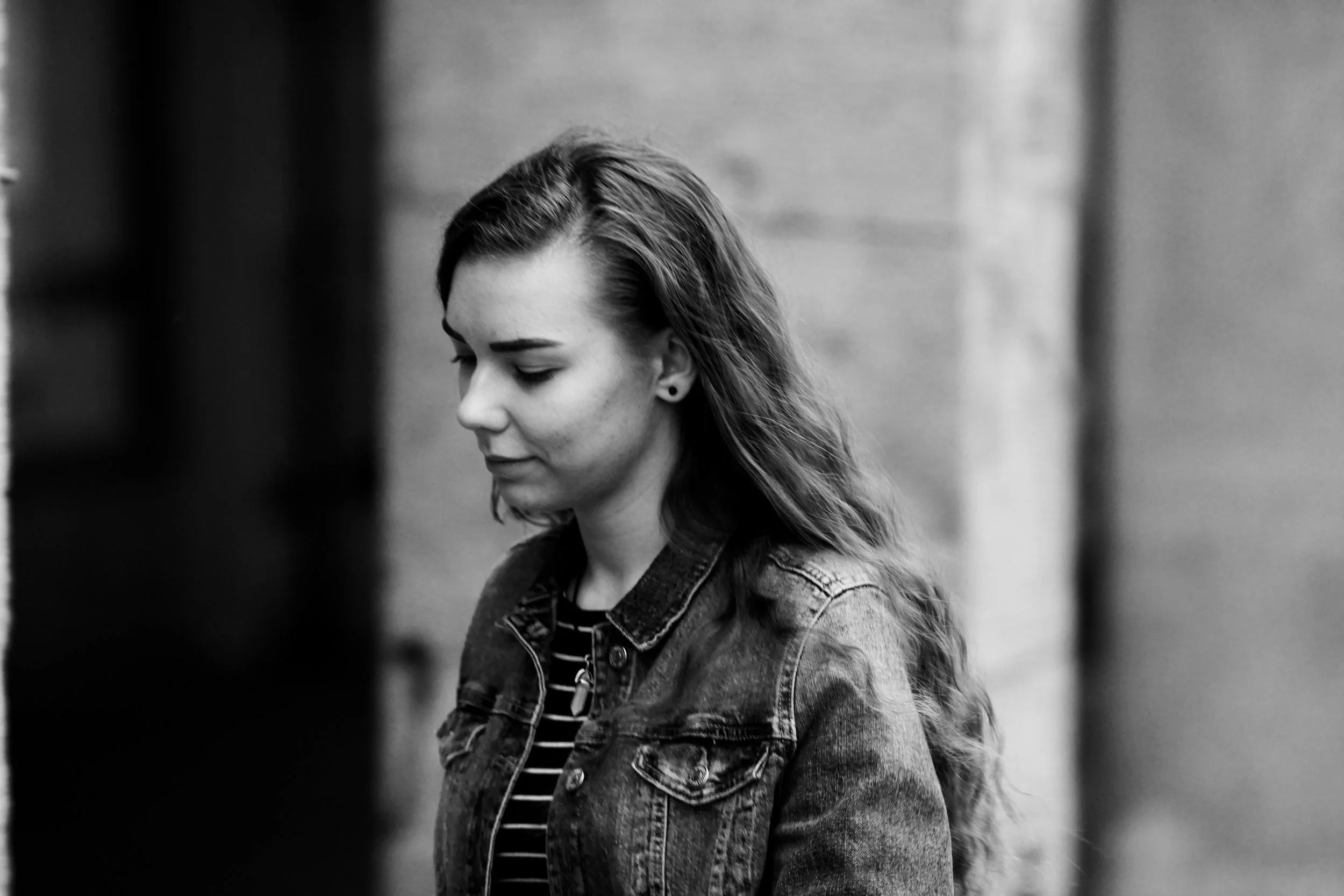 Black and white photo of a young woman with long wavy hair wearing a denim jacket and striped shirt, looking down with her eyes closed.
