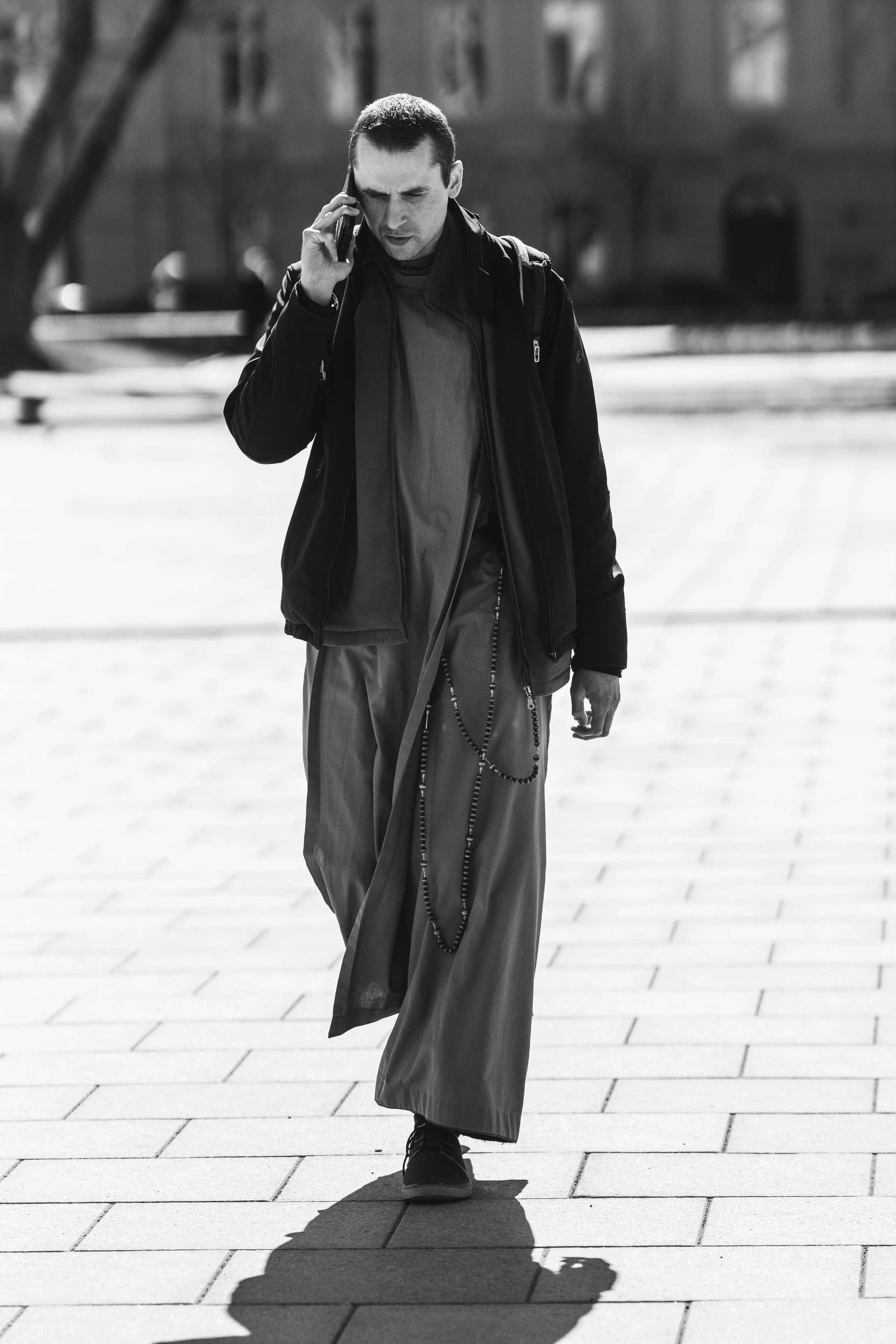 A man walking on a paved sidewalk while talking on his cellphone, dressed in loose pants, a jacket, and sneakers.