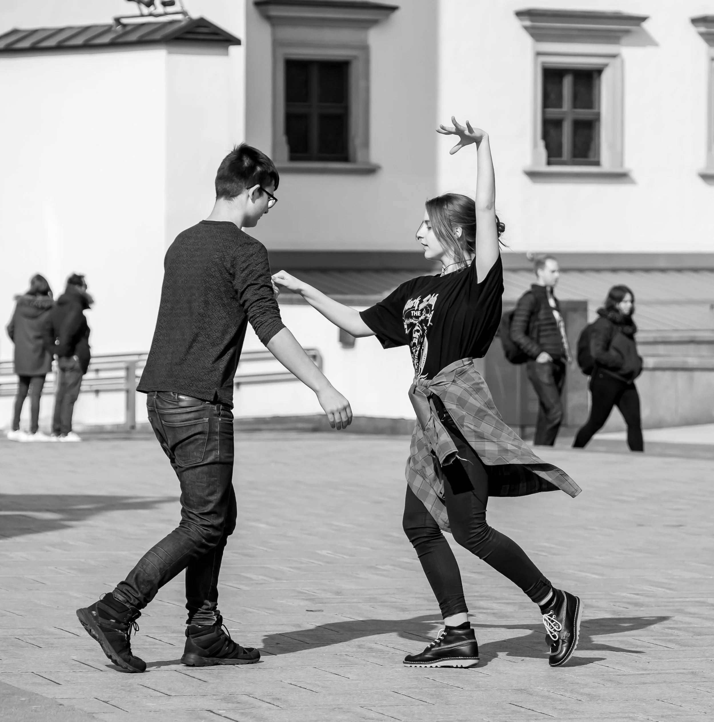 A young man and woman dancing together outdoors in a public square. The woman is mid-dance with her arm raised and wearing a plaid shirt tied around her waist. The man is holding her hand, wearing glasses, and dressed in casual clothing. There are ot
