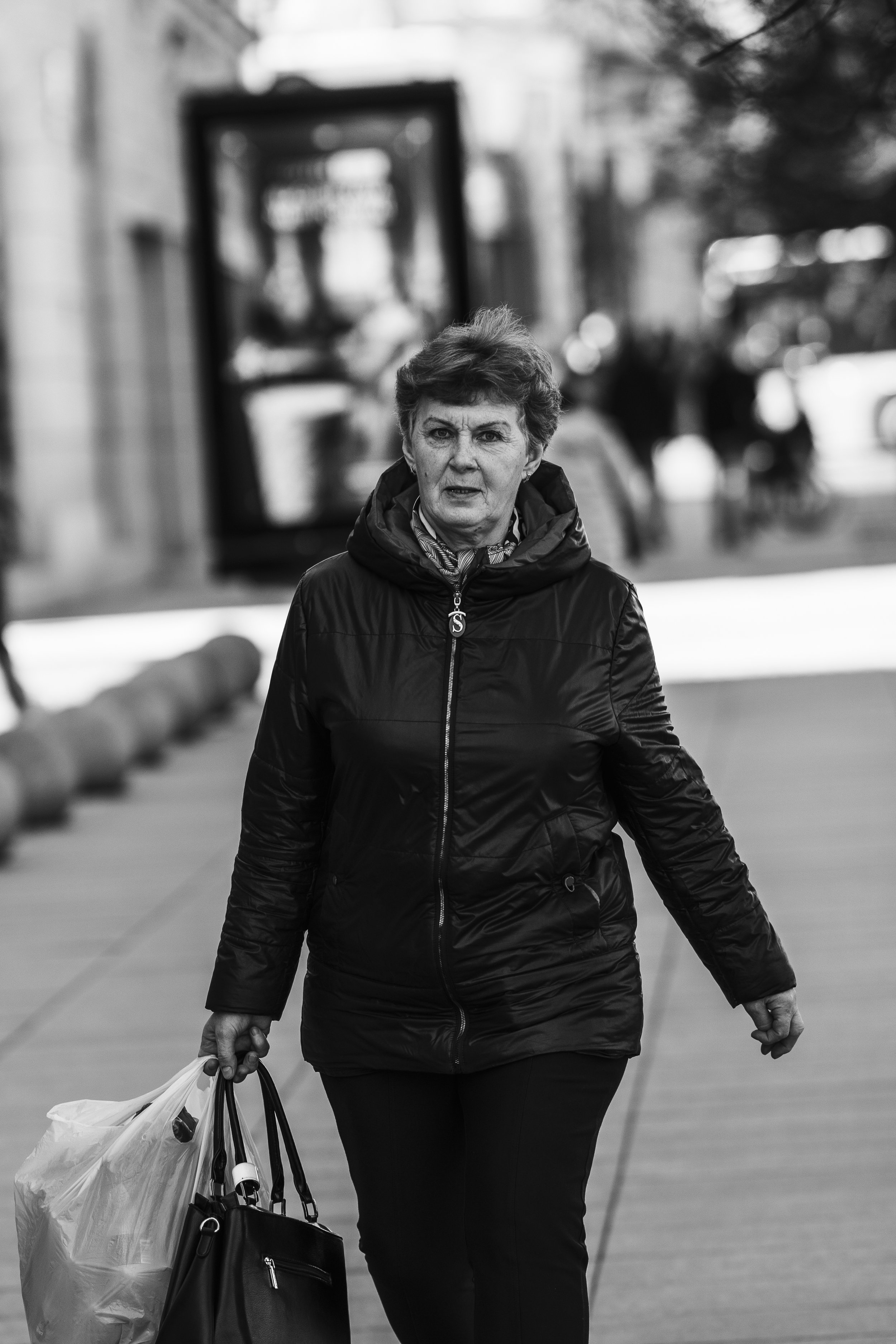 A woman wearing a black jacket and scarf, carrying shopping bags, walking on a city sidewalk.