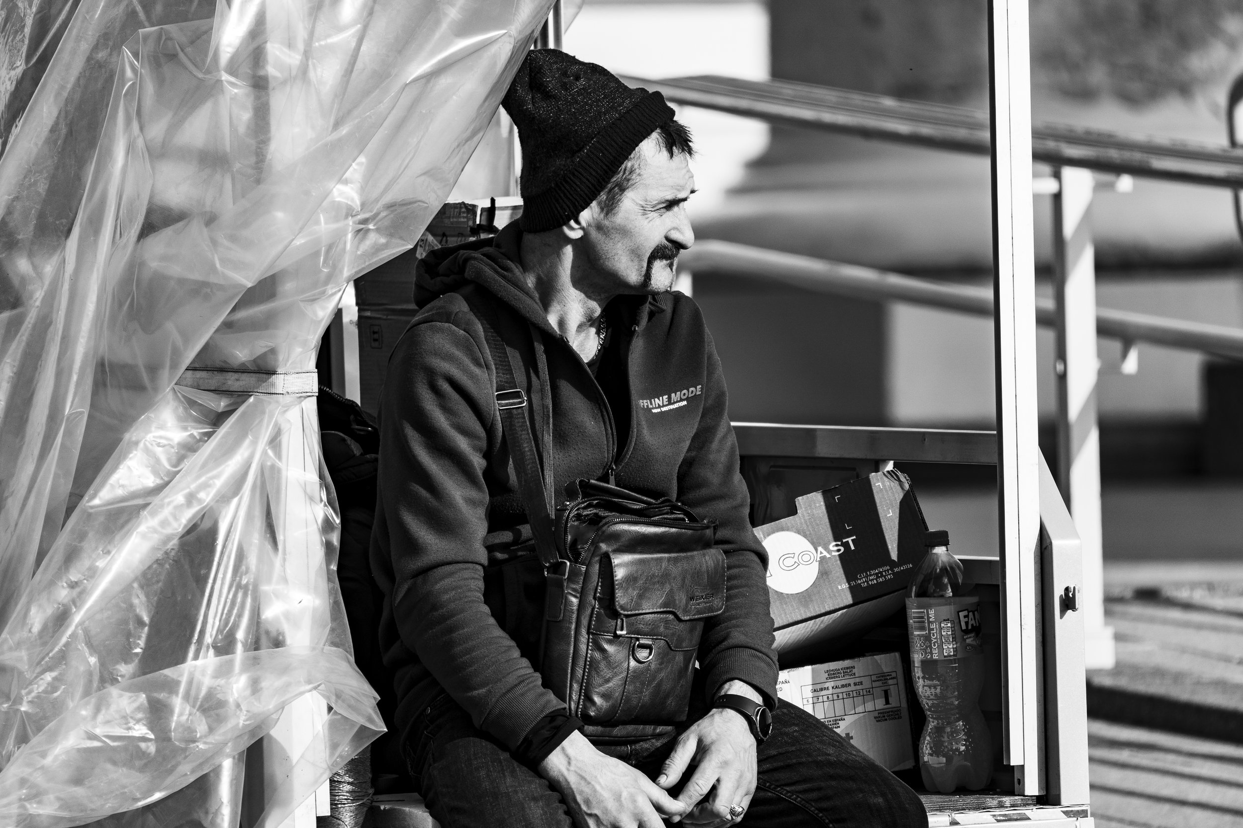 A man sitting on a bench outdoors, wearing a beanie, a hoodie, and holding a leather bag. He appears to be looking into the distance.