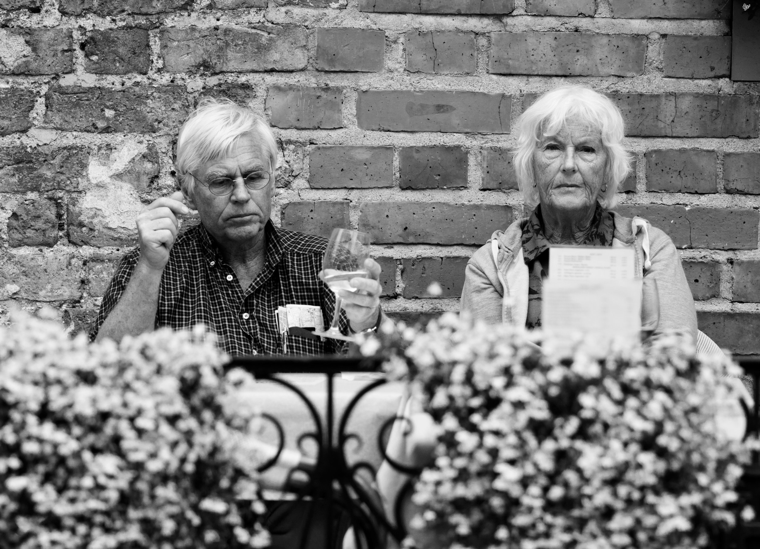An elderly man and woman sitting in front of a brick wall, with a table of flowers in the foreground. The man is holding a wine glass and appears to be listening or contemplating, while the woman looks straight ahead with a serious expression.