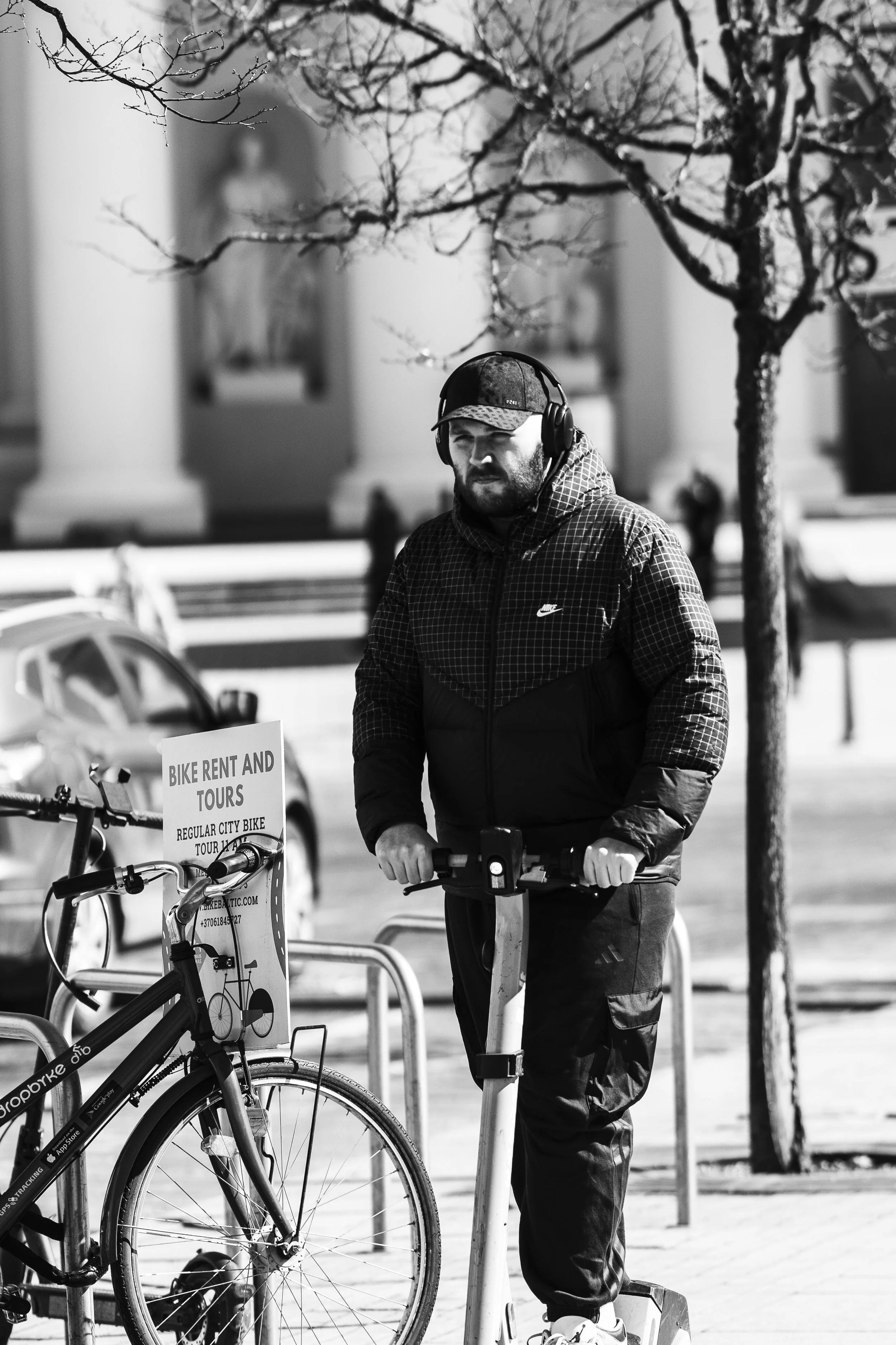 A man wearing a hoodie, jacket, and headphones riding an electric scooter on the city sidewalk, with a bike and a sign offering bike rentals and tours nearby.