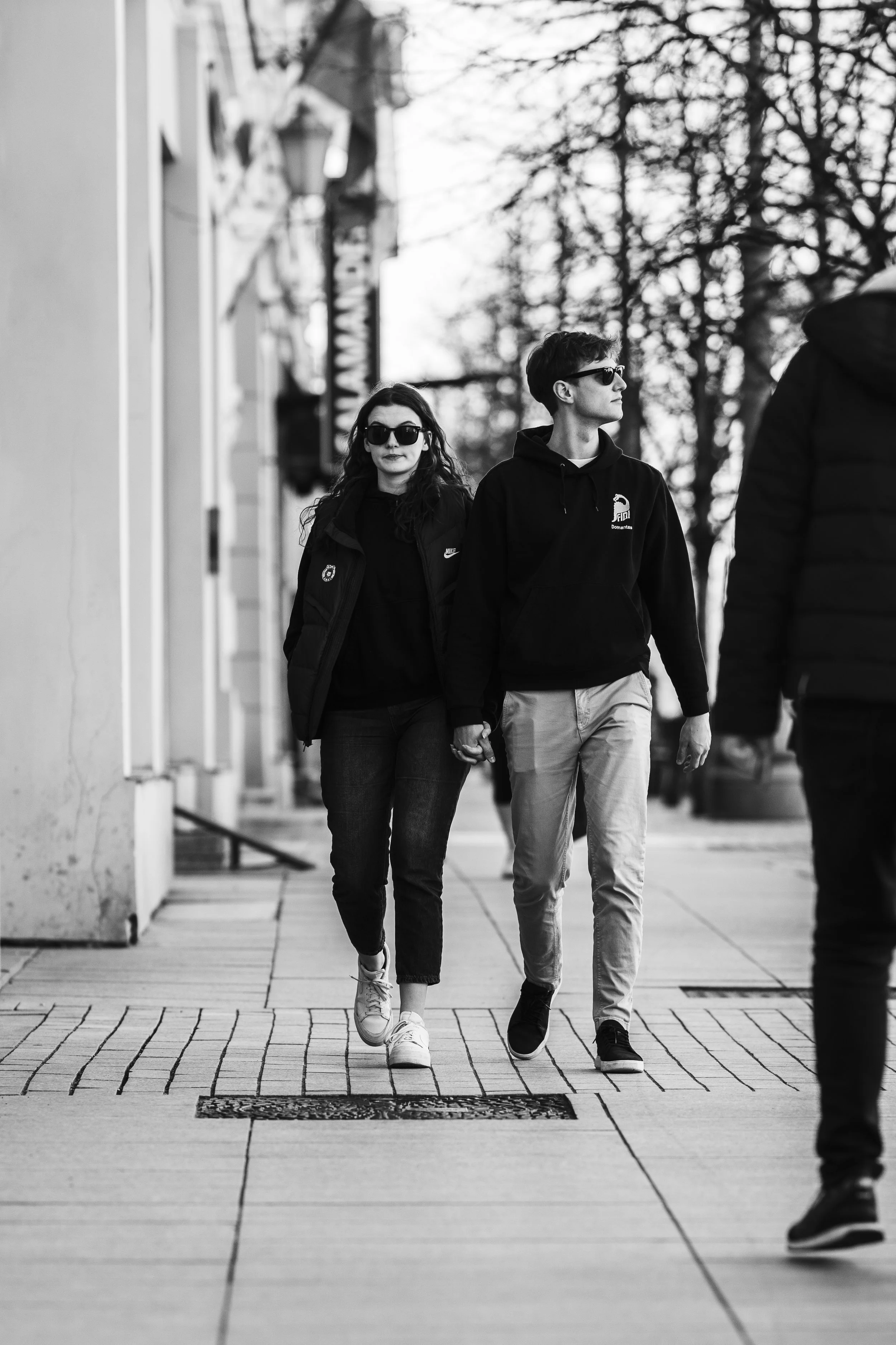 A black and white photo of a young couple walking hand in hand on a city sidewalk, with other pedestrians and trees in the background.