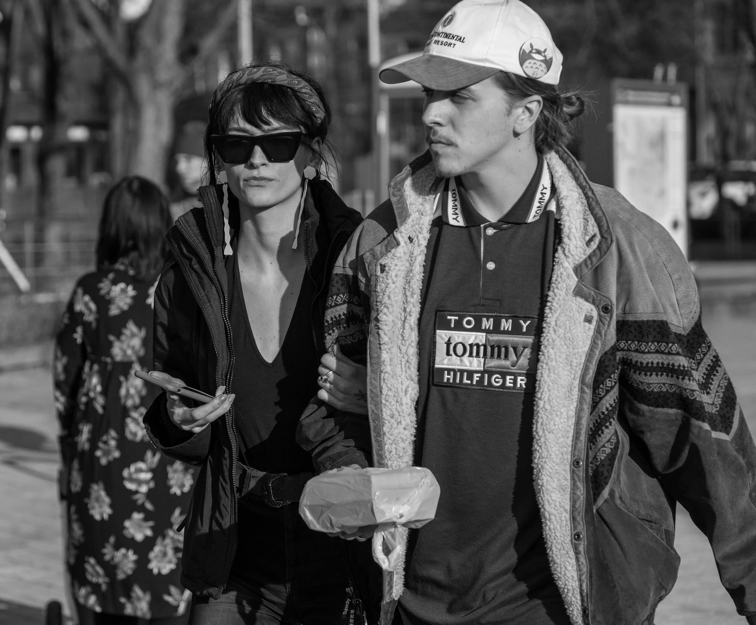 A black-and-white photograph of a man and woman walking outdoors. The woman wears large sunglasses, a headscarf, and a dark jacket, holding a smartphone. The man wears a baseball cap, a patterned jacket with a fleece lining, and a Tommy Hilfiger T-sh