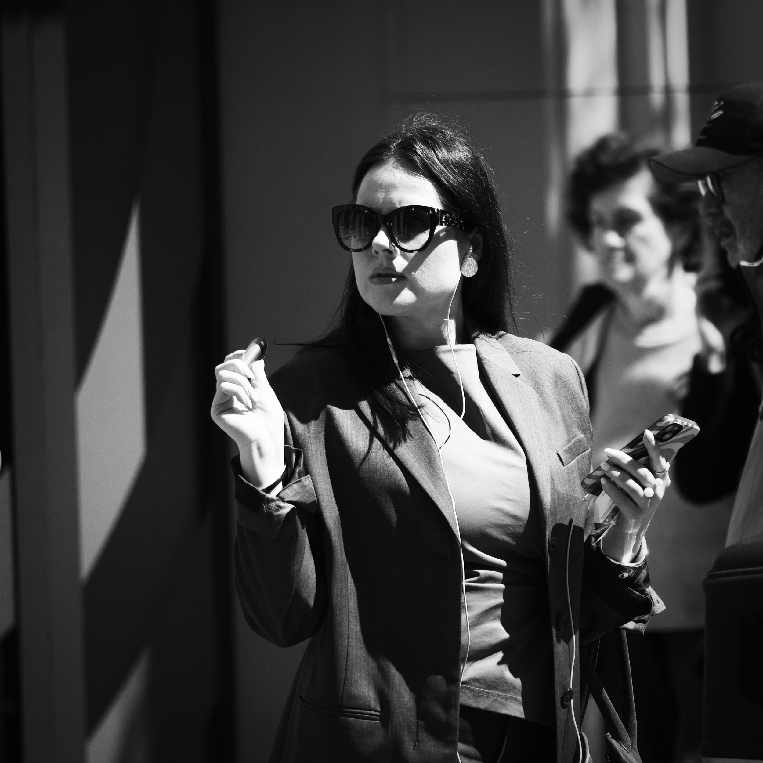 A woman wearing sunglasses and a blazer holding a smartphone and earphones, standing in a busy indoor setting, with two other women in the background.