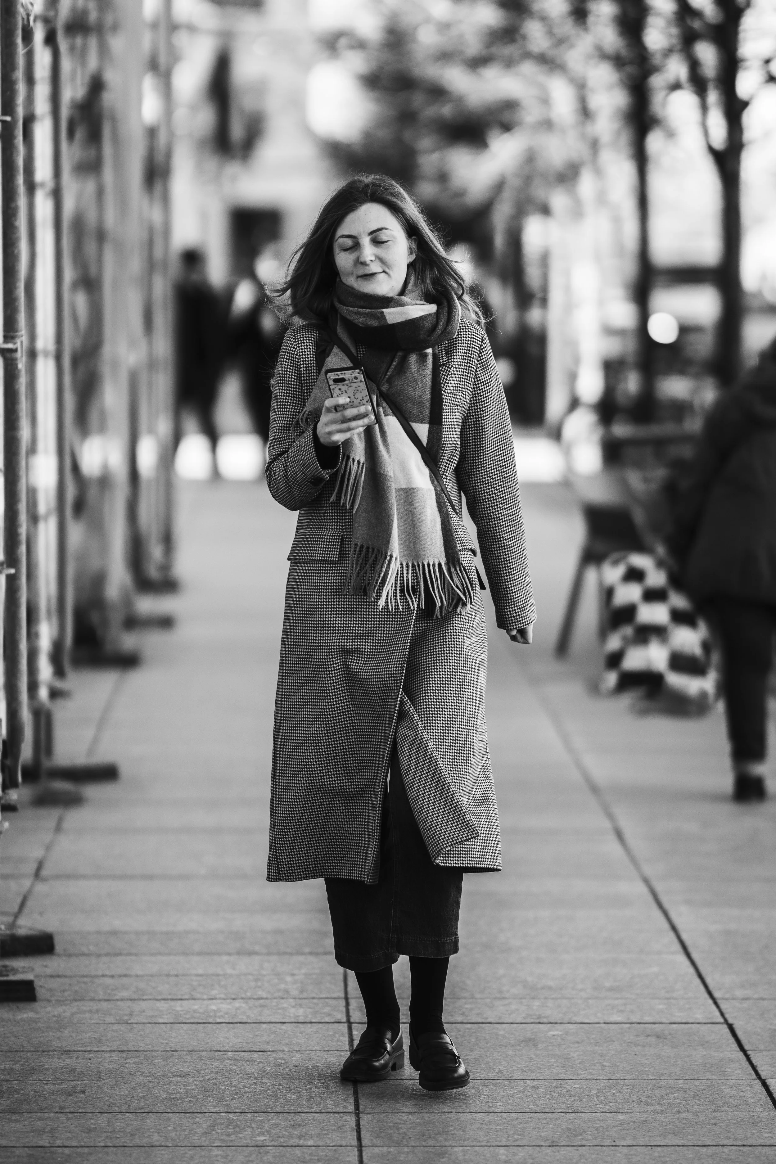 A woman walking on a city sidewalk while looking at her phone, wearing a coat, scarf, and boots in an urban setting.
