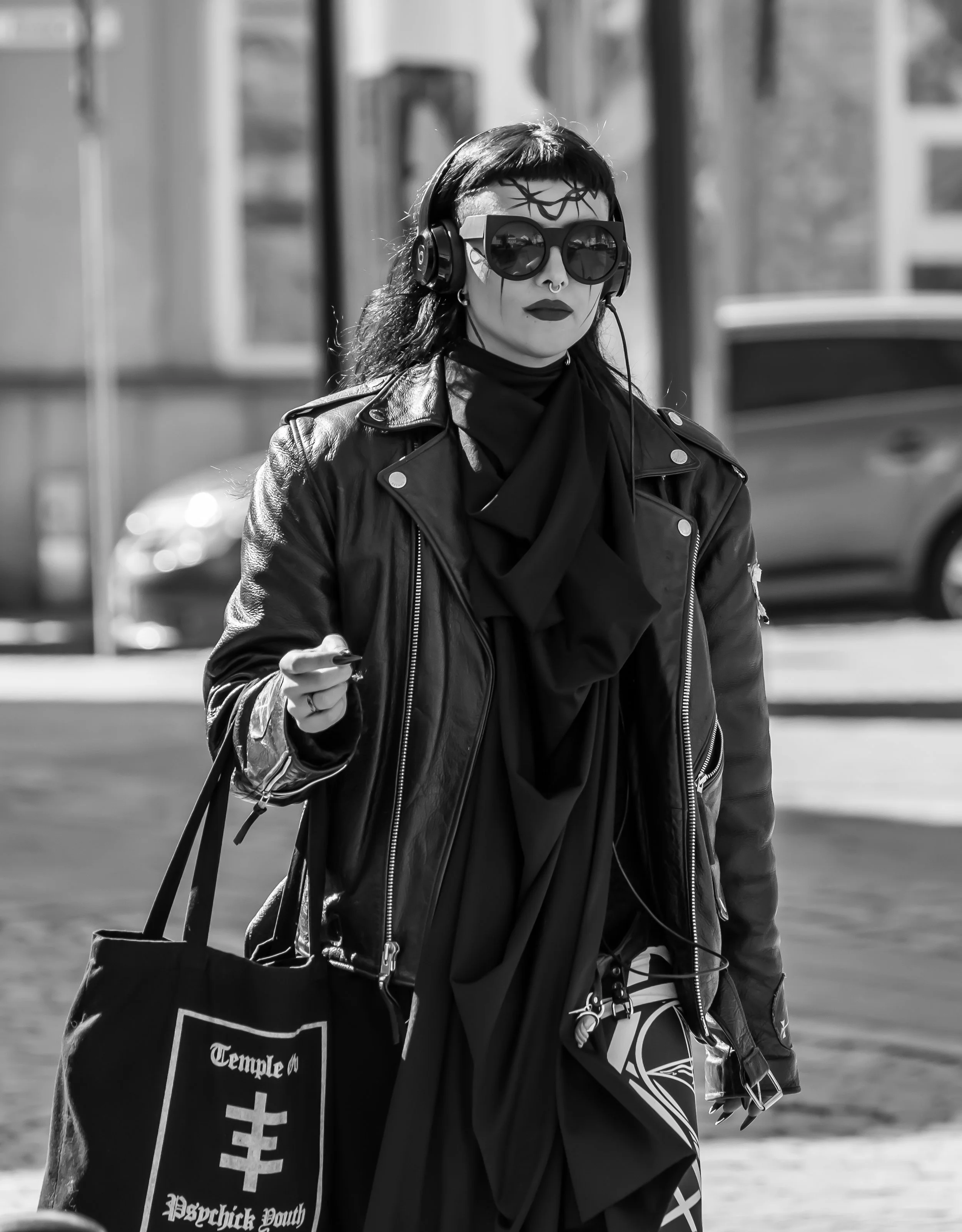 A woman with dark hair and tattoos, wearing large sunglasses, headphones, a leather jacket, and a dark scarf, walking outdoors while carrying a bag with 'Temple of Decadence Youth' written on it.