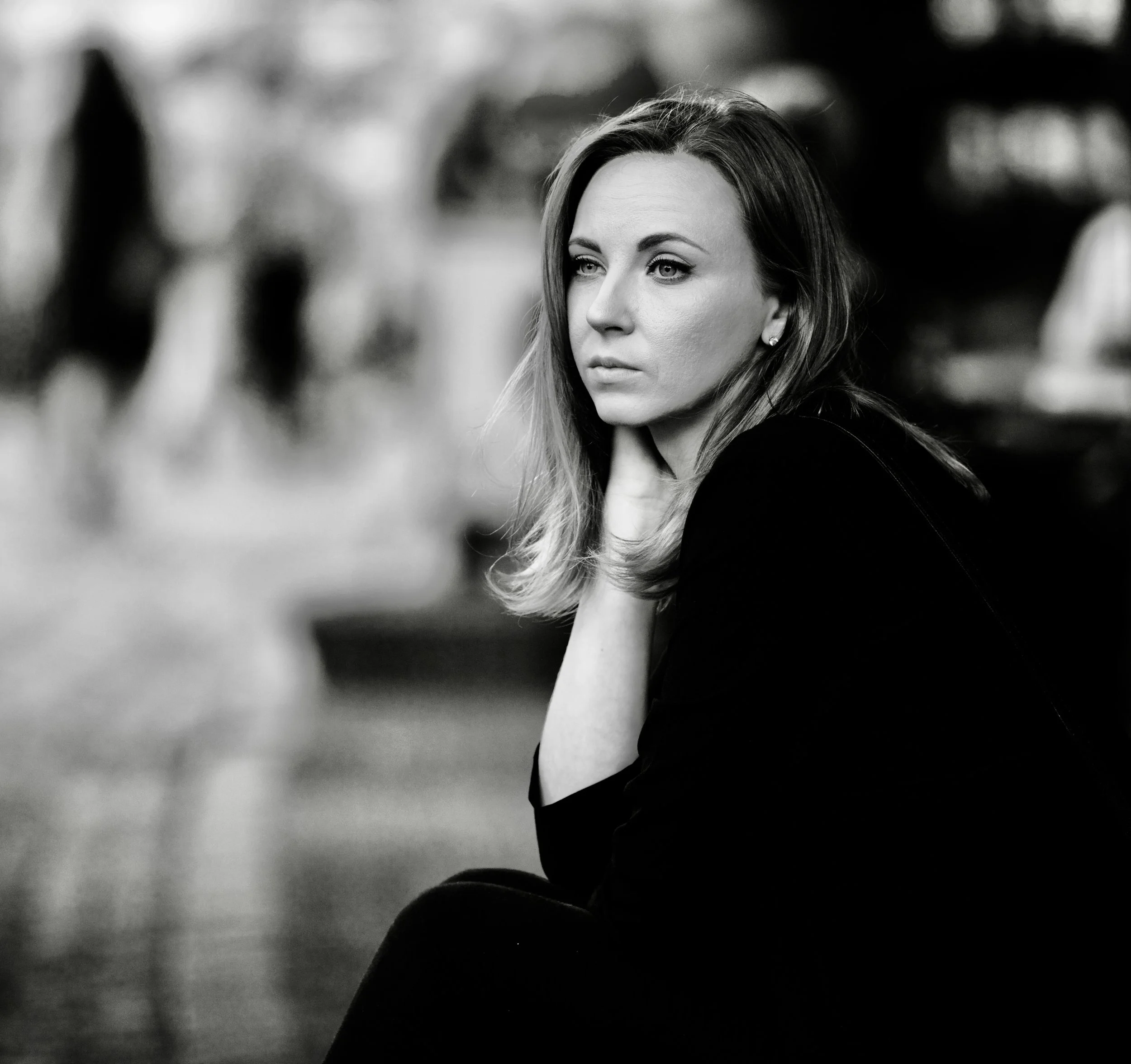 A black and white photograph of a woman with light shoulder-length hair, sitting outdoors with a contemplative expression, resting her chin on her hand, with blurred background of people and trees.