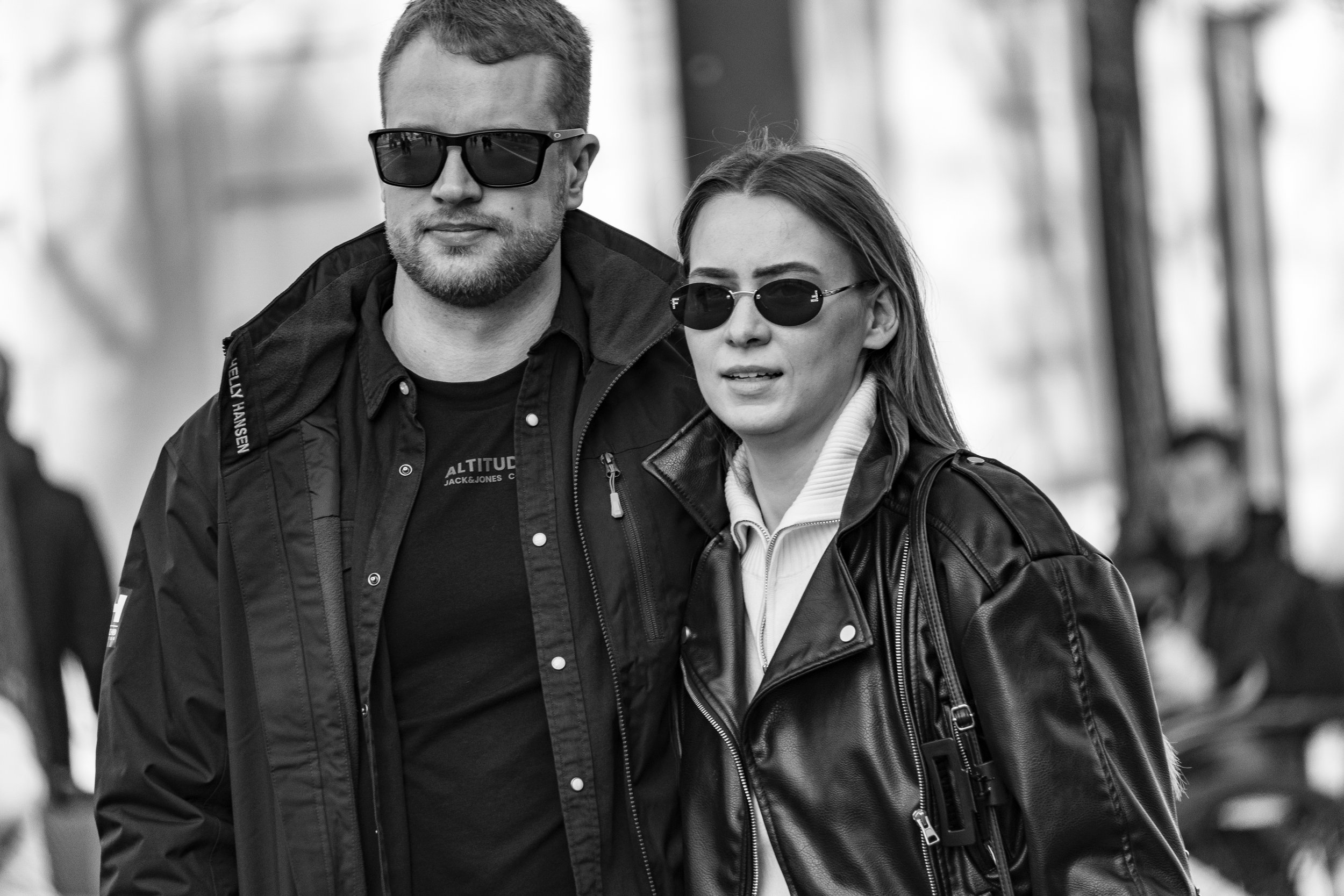 A man and woman wearing sunglasses walking outdoors, dressed in casual jackets.