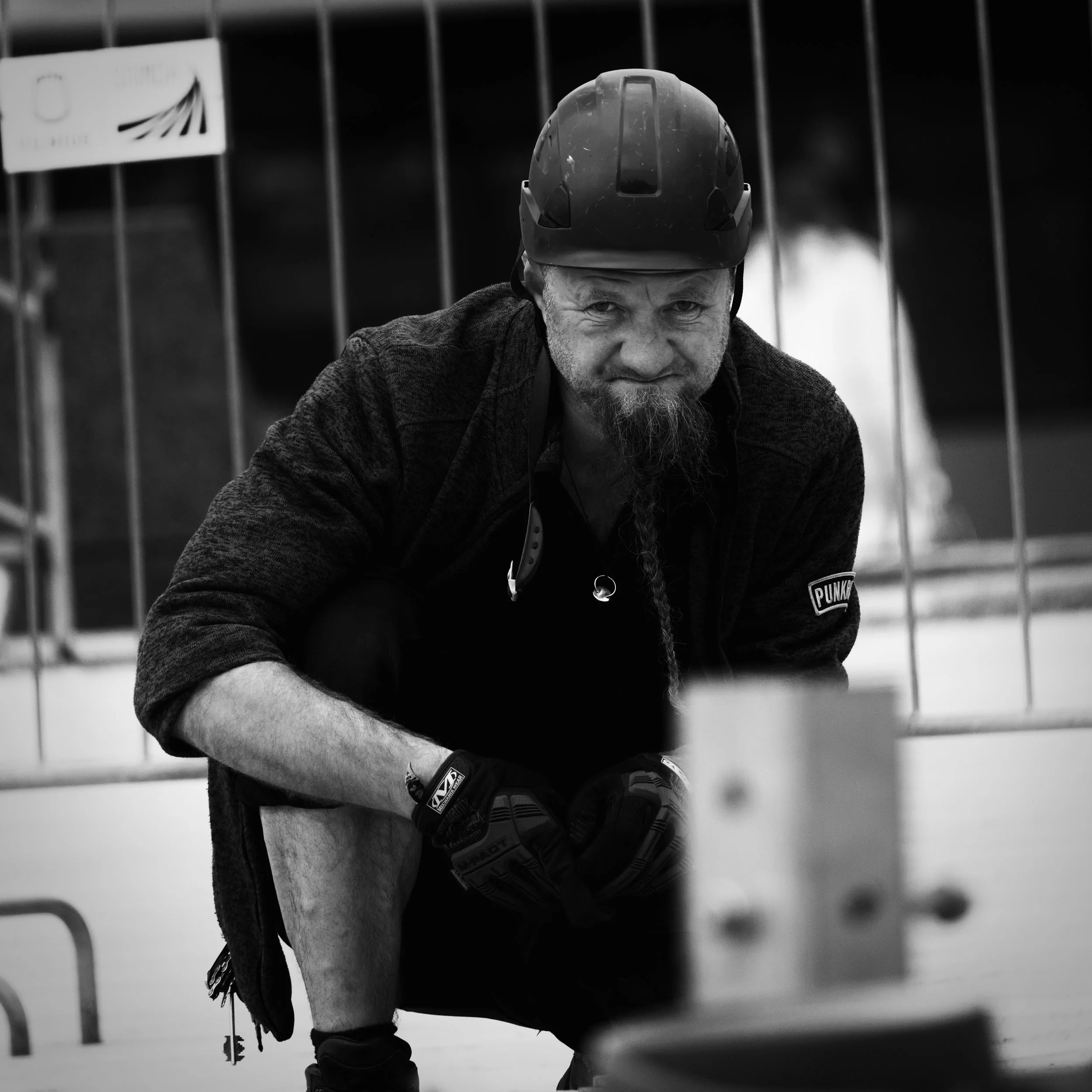 A man with a beard and mustache wearing a safety helmet, gloves, and a jacket, crouching indoors behind a metal barrier in black and white.