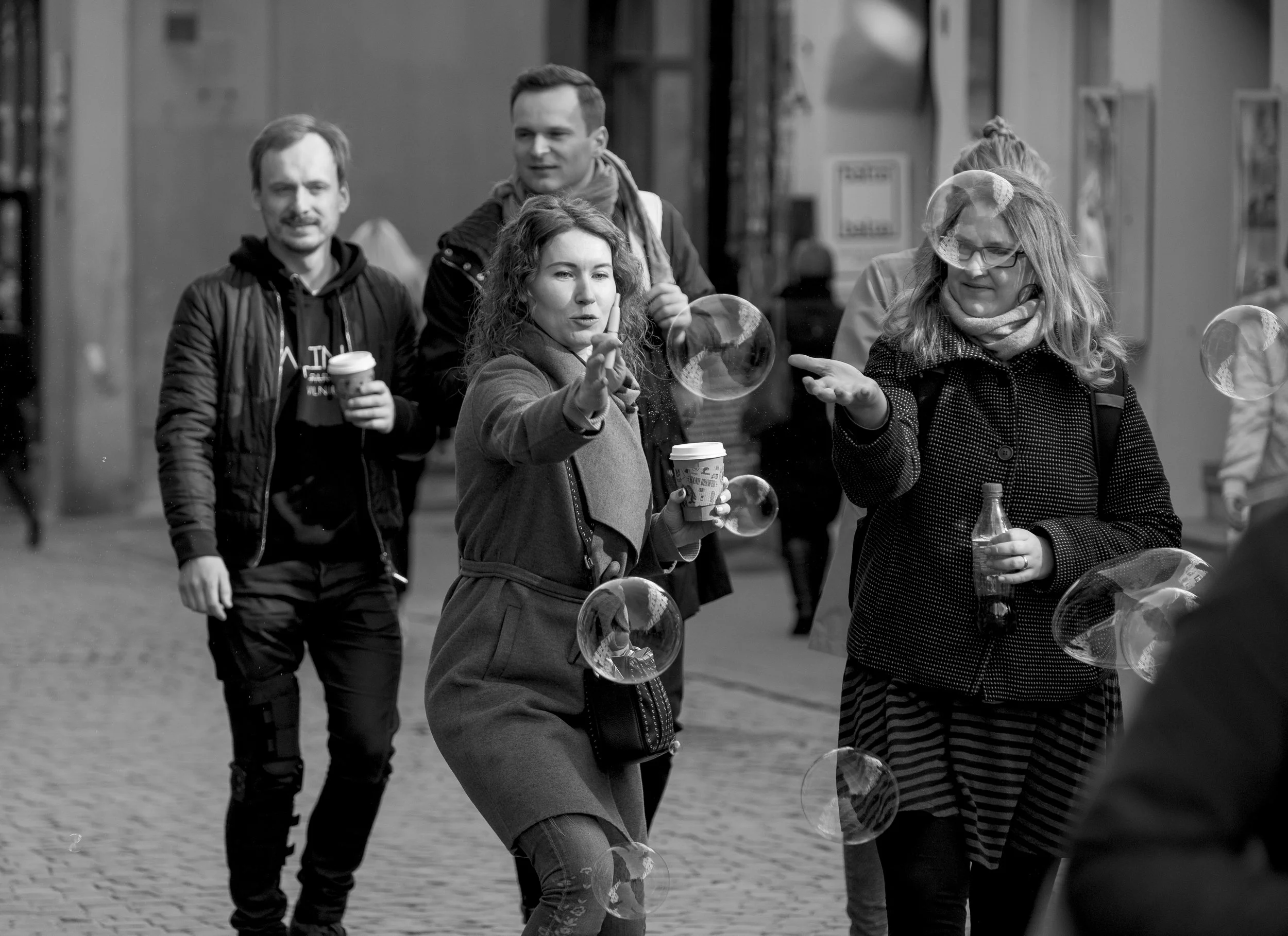 People walking on a city street, with two women in the foreground blowing bubbles, and two men behind them holding cups of drinks.