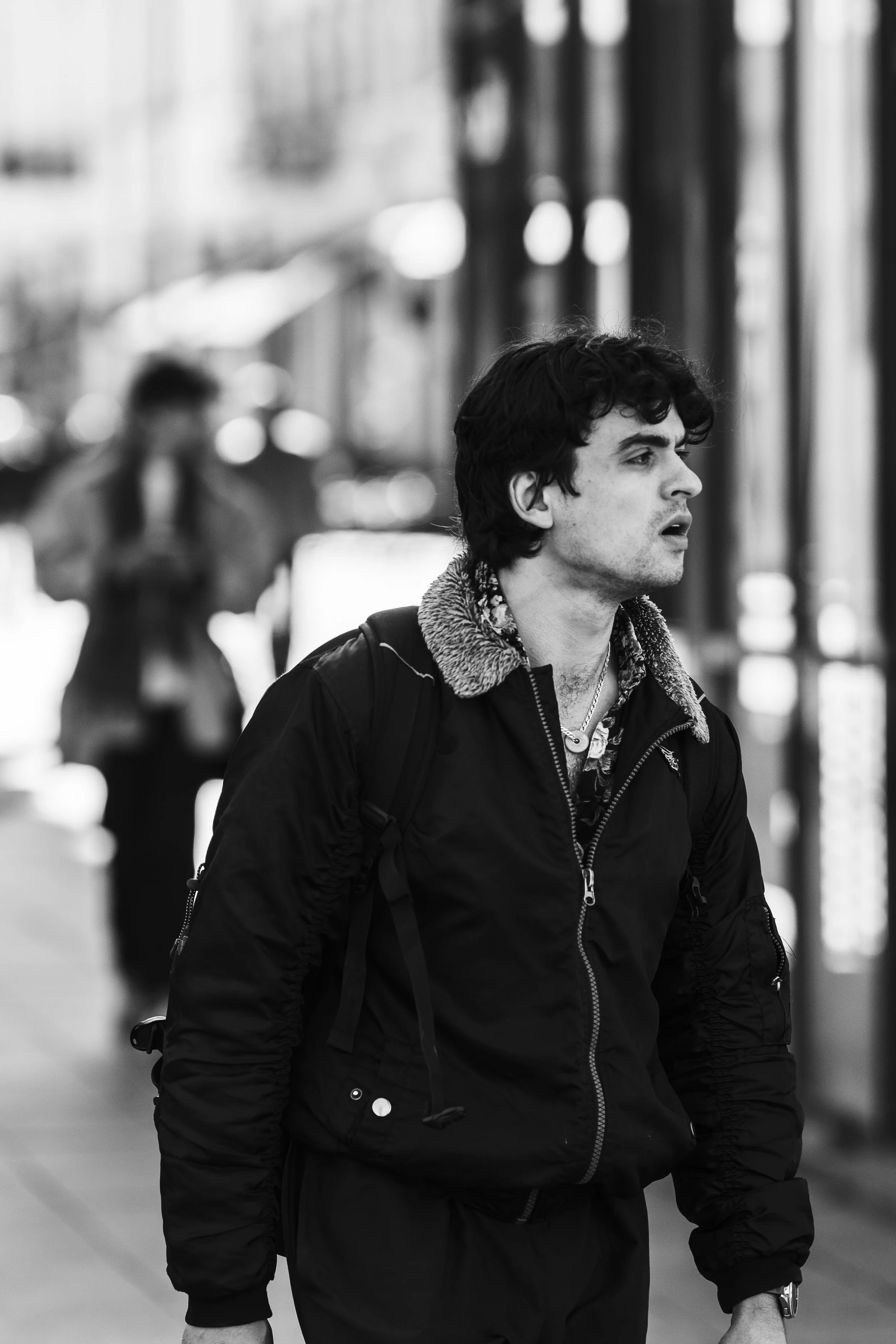 Black and white photo of a young man with dark wavy hair walking on a city street, wearing a jacket with a furry collar and carrying a backpack. A woman is walking behind him, looking at her phone.