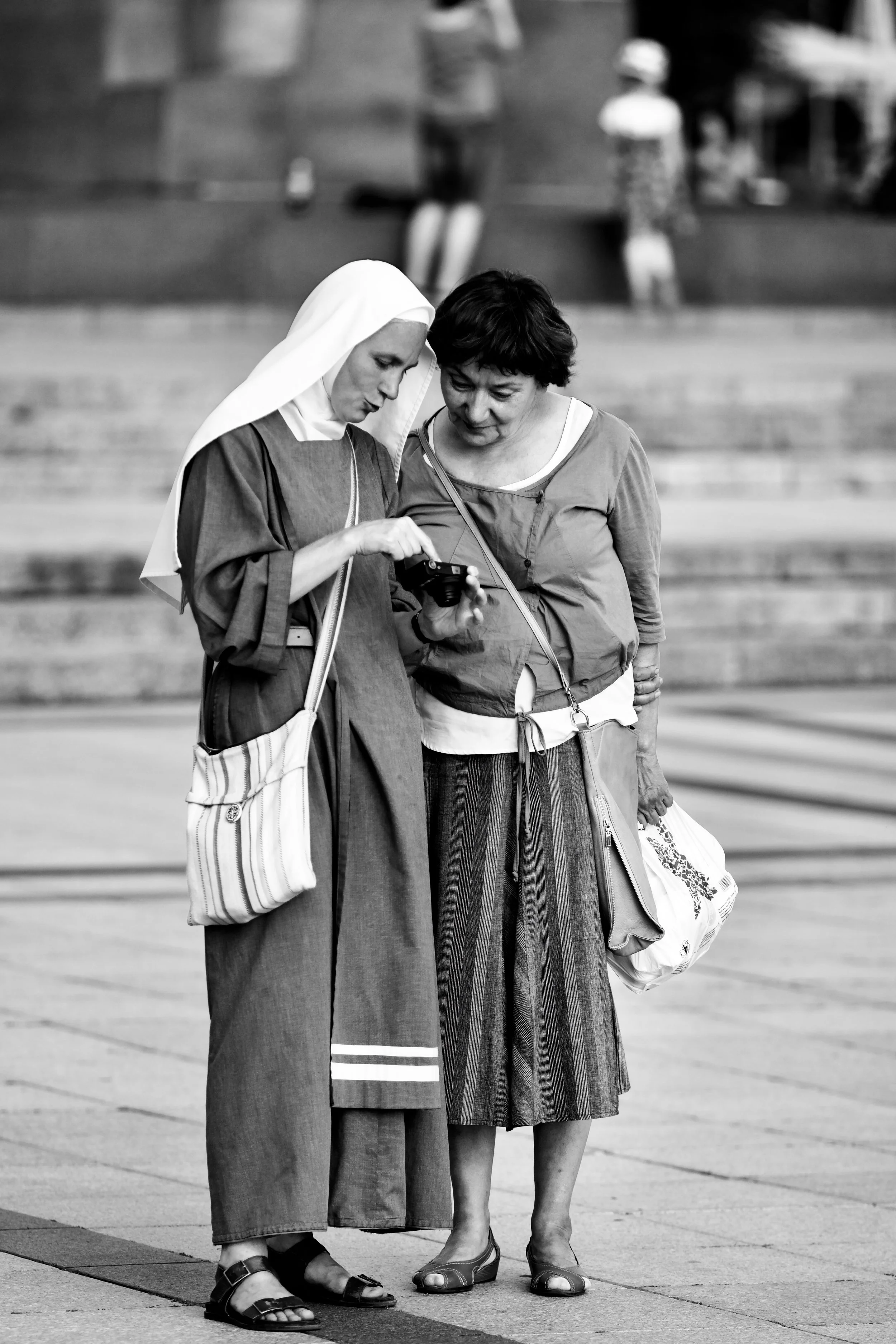 Two women standing close together, looking at a camera or phone, in an outdoor urban setting with stairs and other figures in the background.