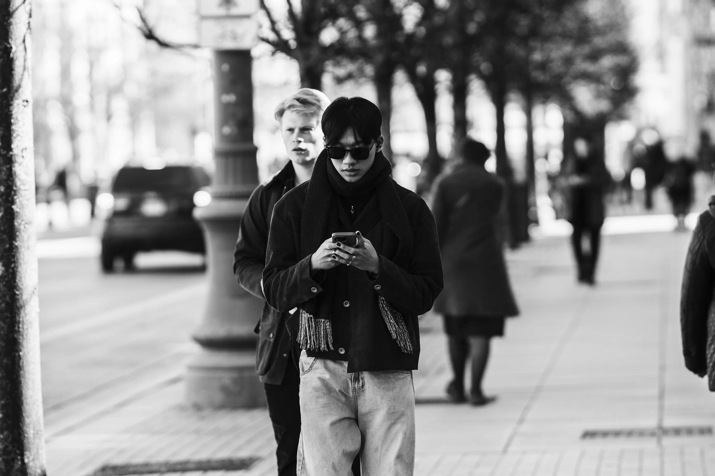 Black and white photo of a woman with short hair, wearing sunglasses and a scarf, looking at her phone on a city sidewalk. Behind her, a man with light hair looks in her direction. Other pedestrians and blurred cars are visible in the background.