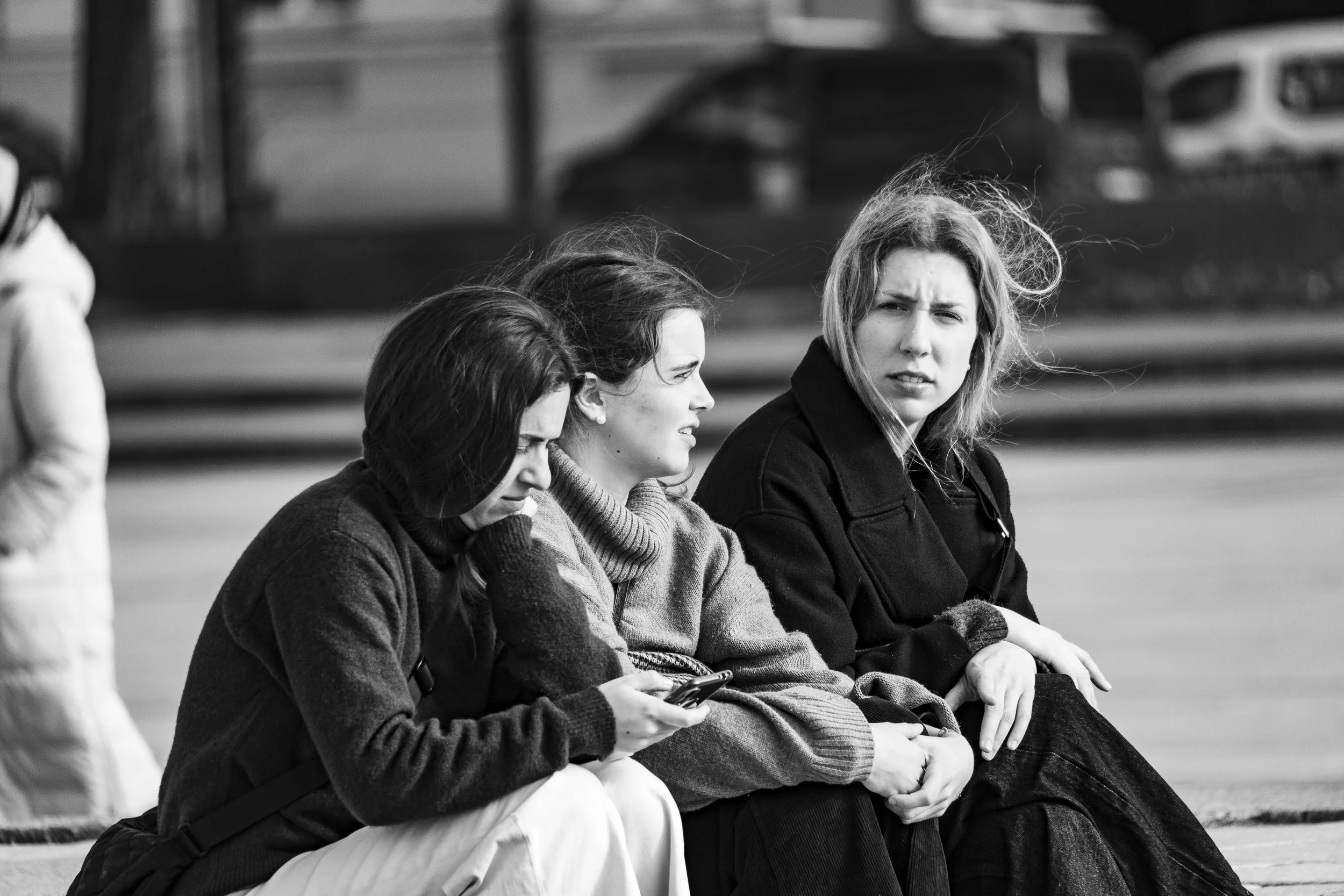 Three women sit on a bench outdoors, with two of them looking at their phones and the third looking off into the distance with a concerned expression, in black and white.