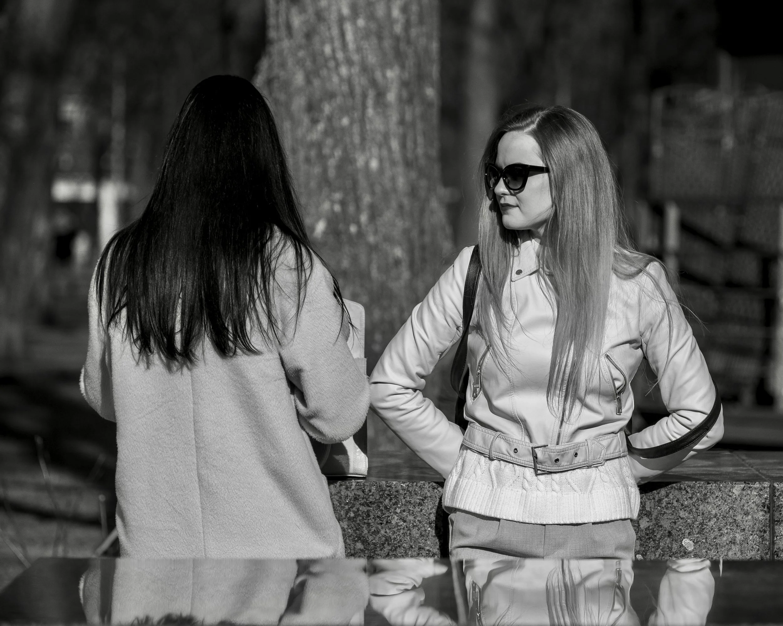 Two women talking outdoors, one with sunglasses and long hair, the other with her back to the camera, in black and white.
