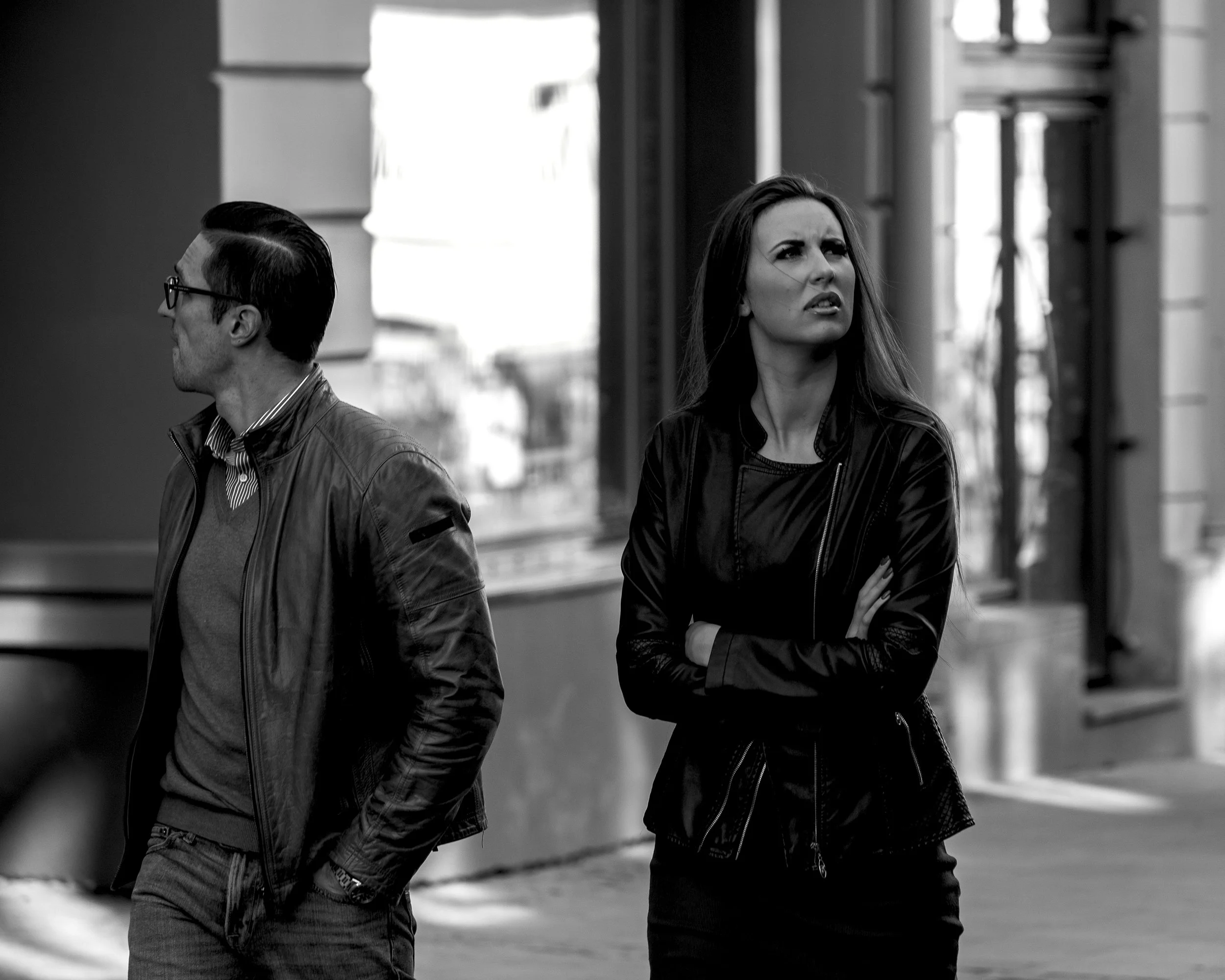 A black and white photo of a man and a woman standing on a sidewalk. The man on the left is wearing glasses, a leather jacket, and looking away. The woman on the right is with crossed arms, looking confused or annoyed, wearing a leather jacket.