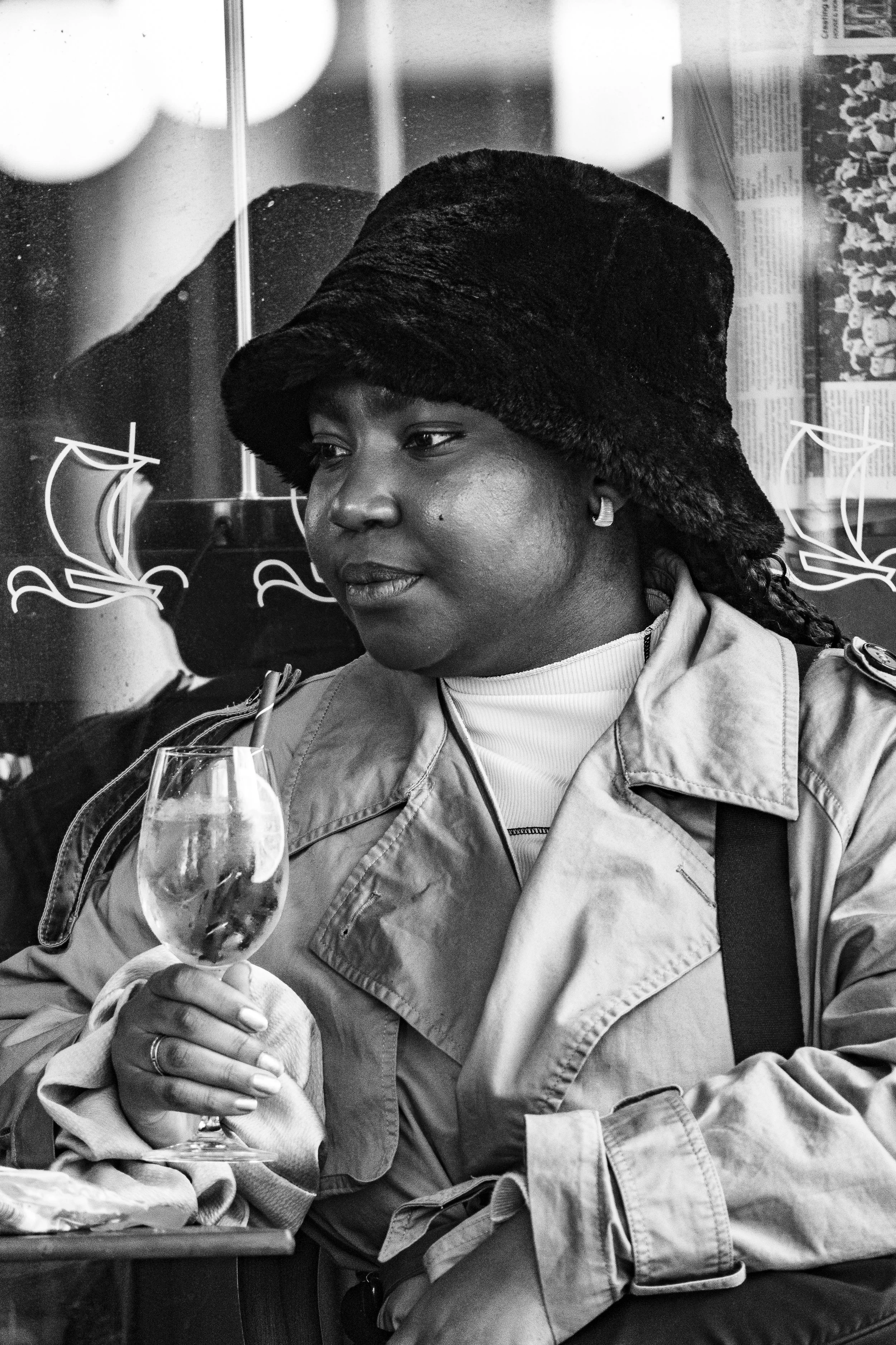 A woman with dark skin, wearing a large hat and a trench coat, holding a wine glass, seated indoors with a reflective glass wall behind her.