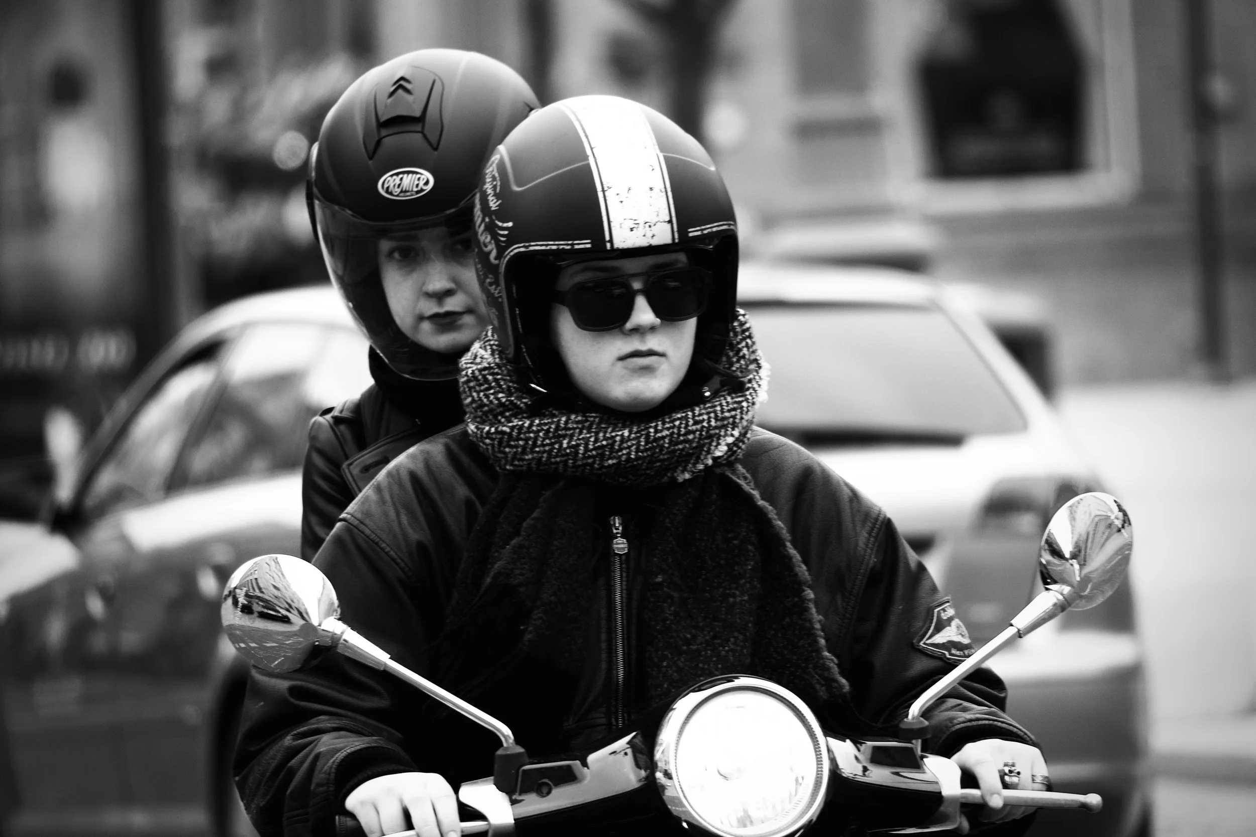 Two women riding a motorcycle in an urban street, both wearing helmets and jackets, one with sunglasses and a scarf, captured in black and white.