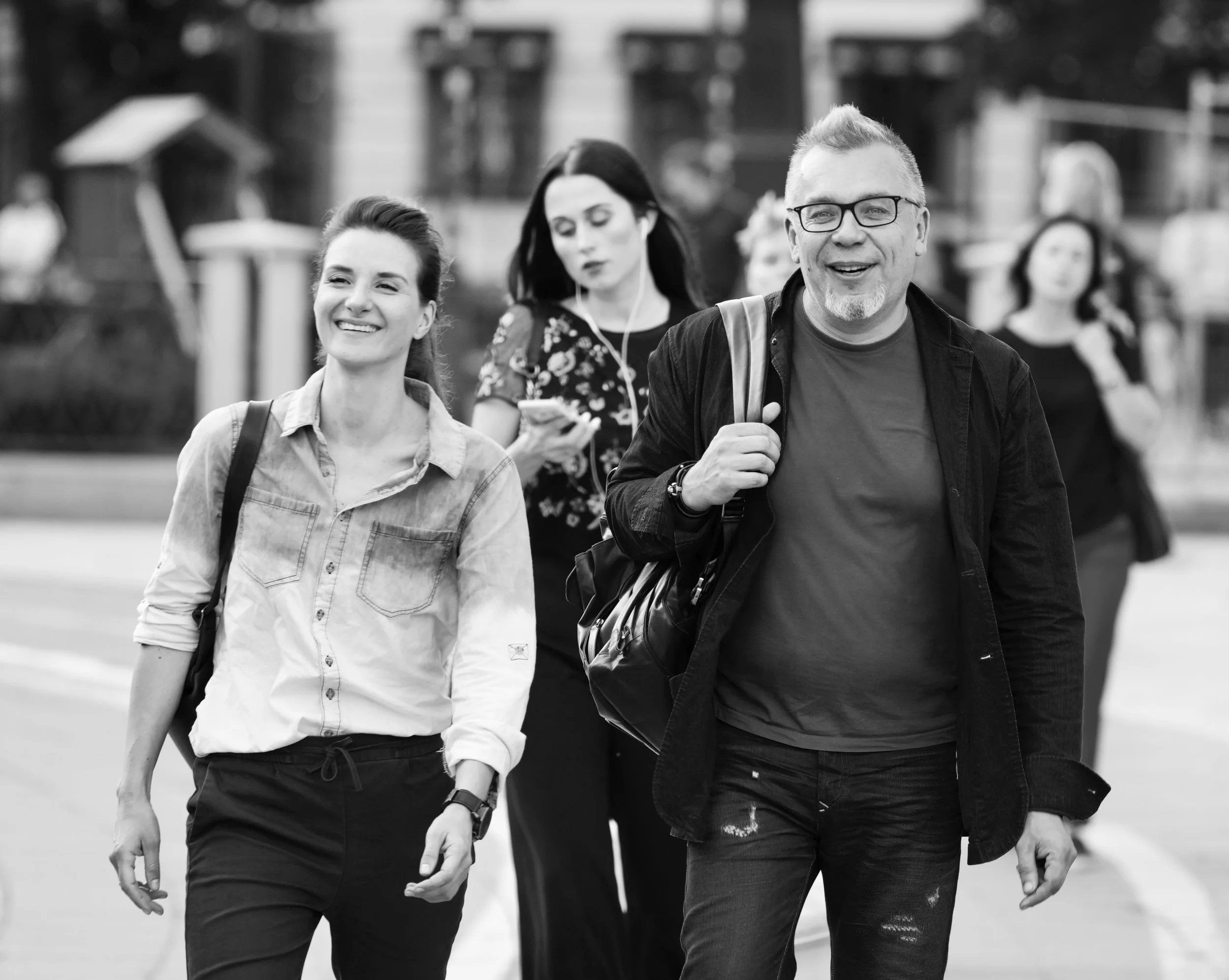 A group of people walking outdoors in an urban area, with a woman smiling in the foreground and a man wearing glasses and carrying a backpack. Other women are visible in the background, one looking at her phone.