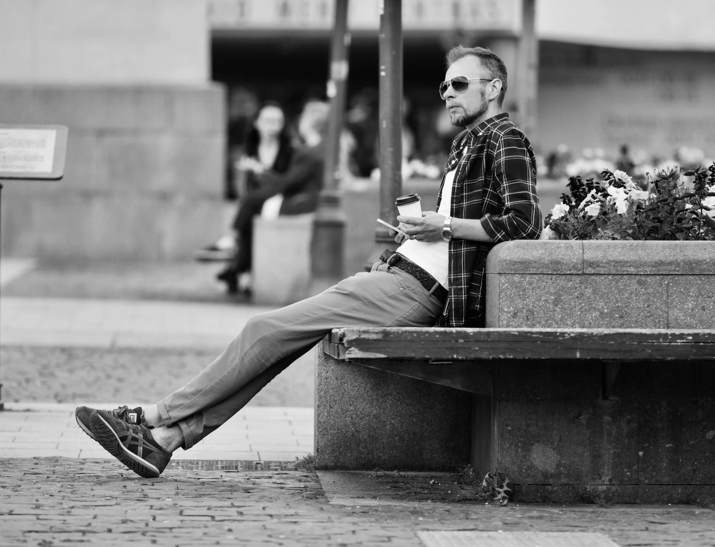 A man sitting on a park bench holding a coffee cup and a smartphone, wearing sunglasses and a plaid shirt, with people blurred in the background.