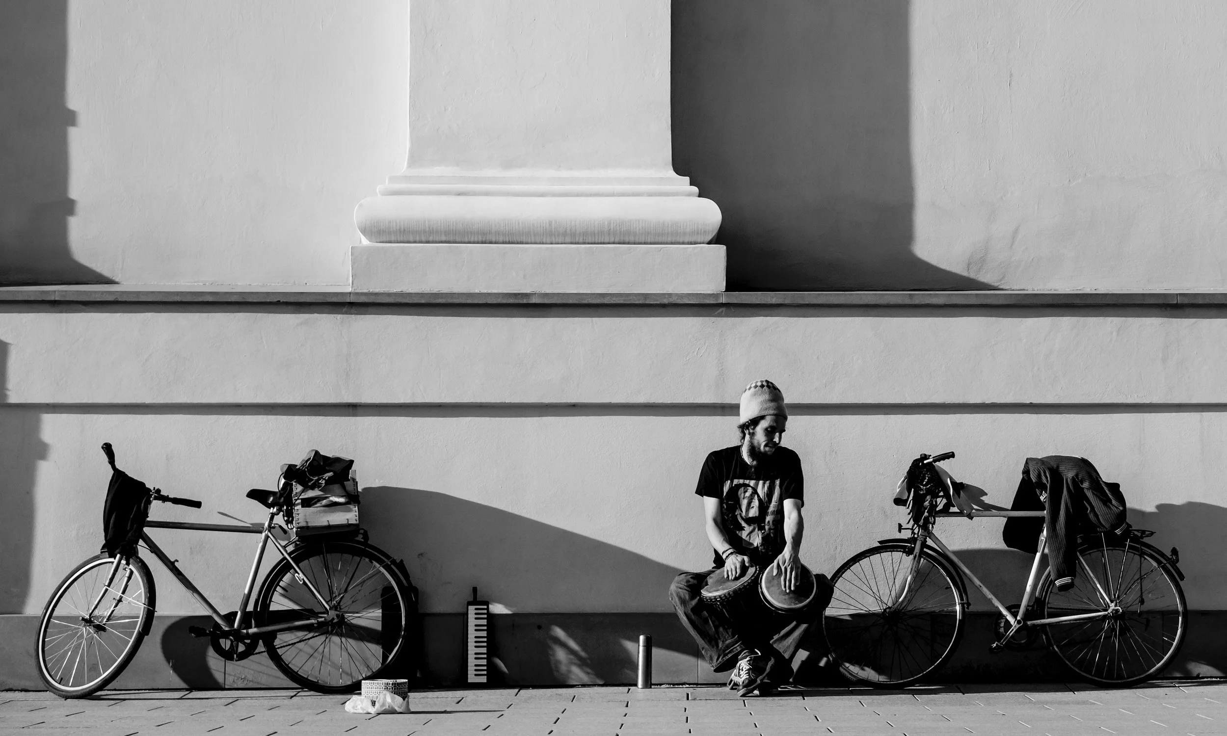 A man wearing a beanie and black t-shirt playing drums while sitting on the ground, flanked by two bicycles, with shadows cast on the wall behind him.