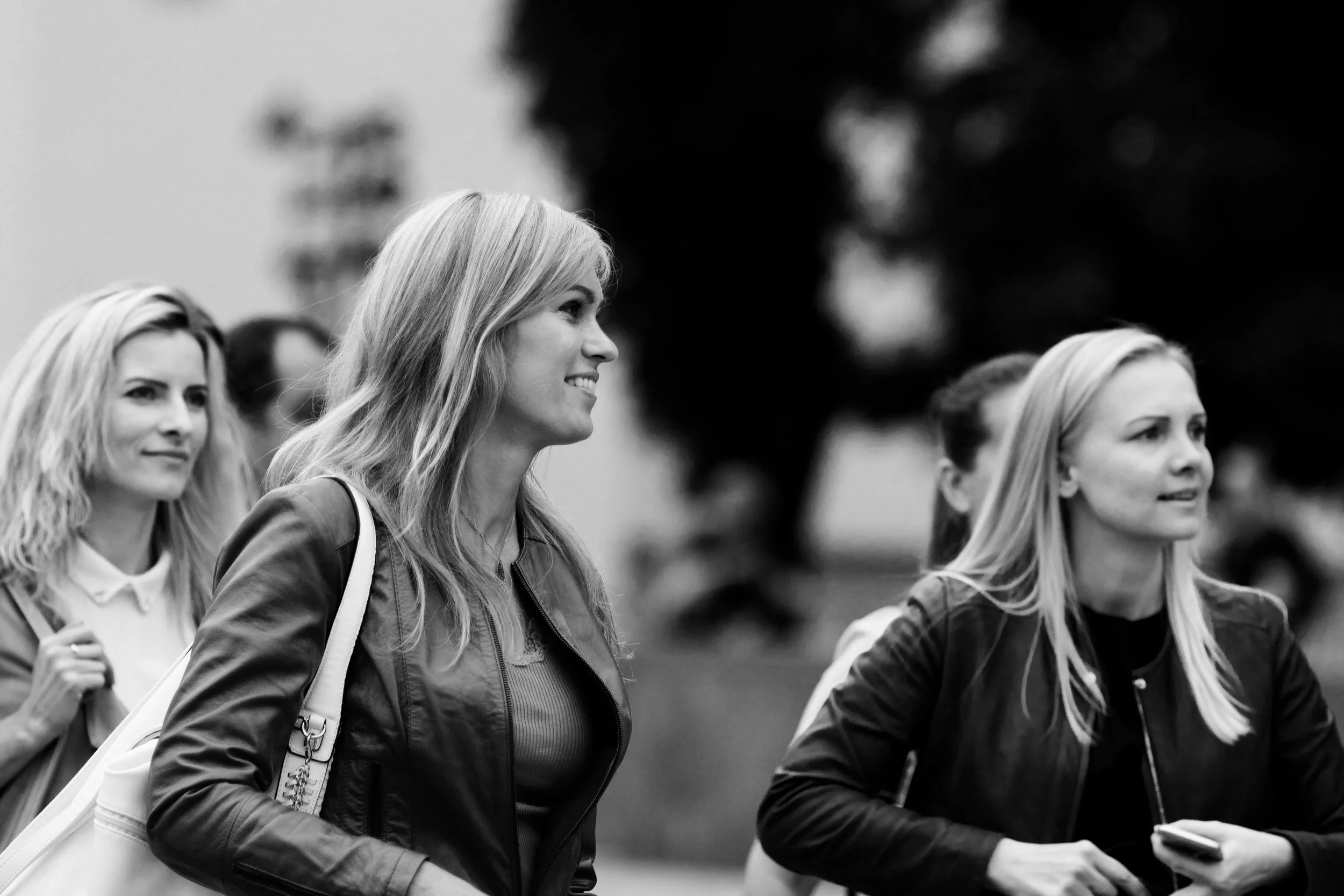 Three women walking outdoors, smiling, with one woman holding a smartphone, in the background with blurred trees.