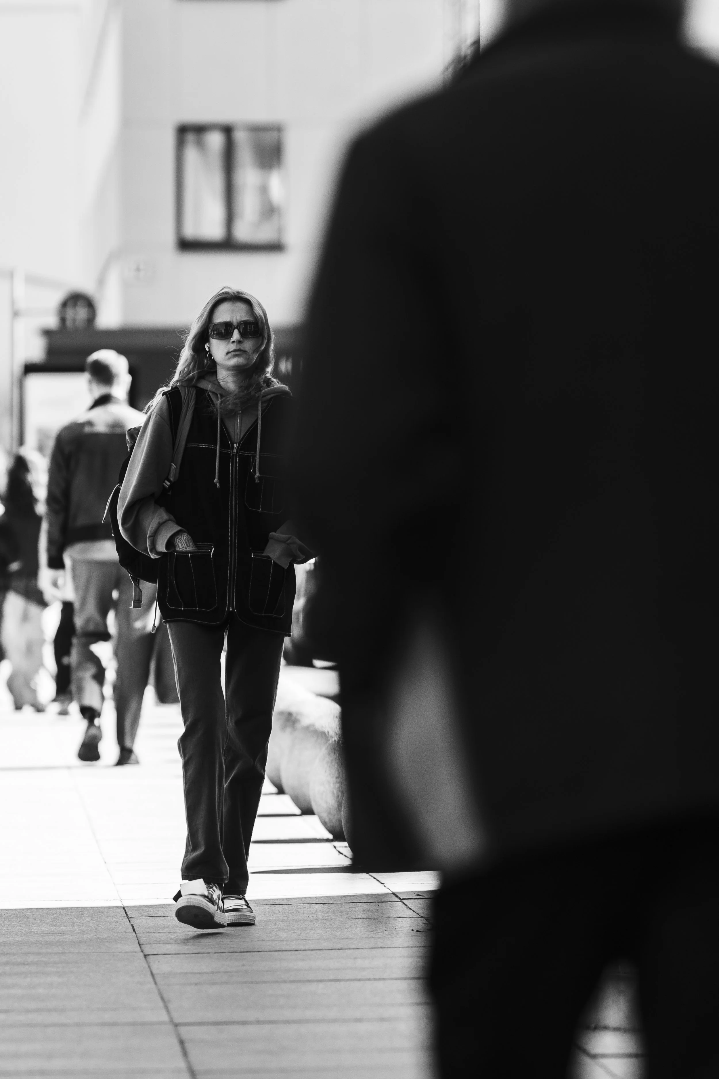 A woman wearing sunglasses and casual clothing walking on a city sidewalk among other pedestrians in black and white photograph.