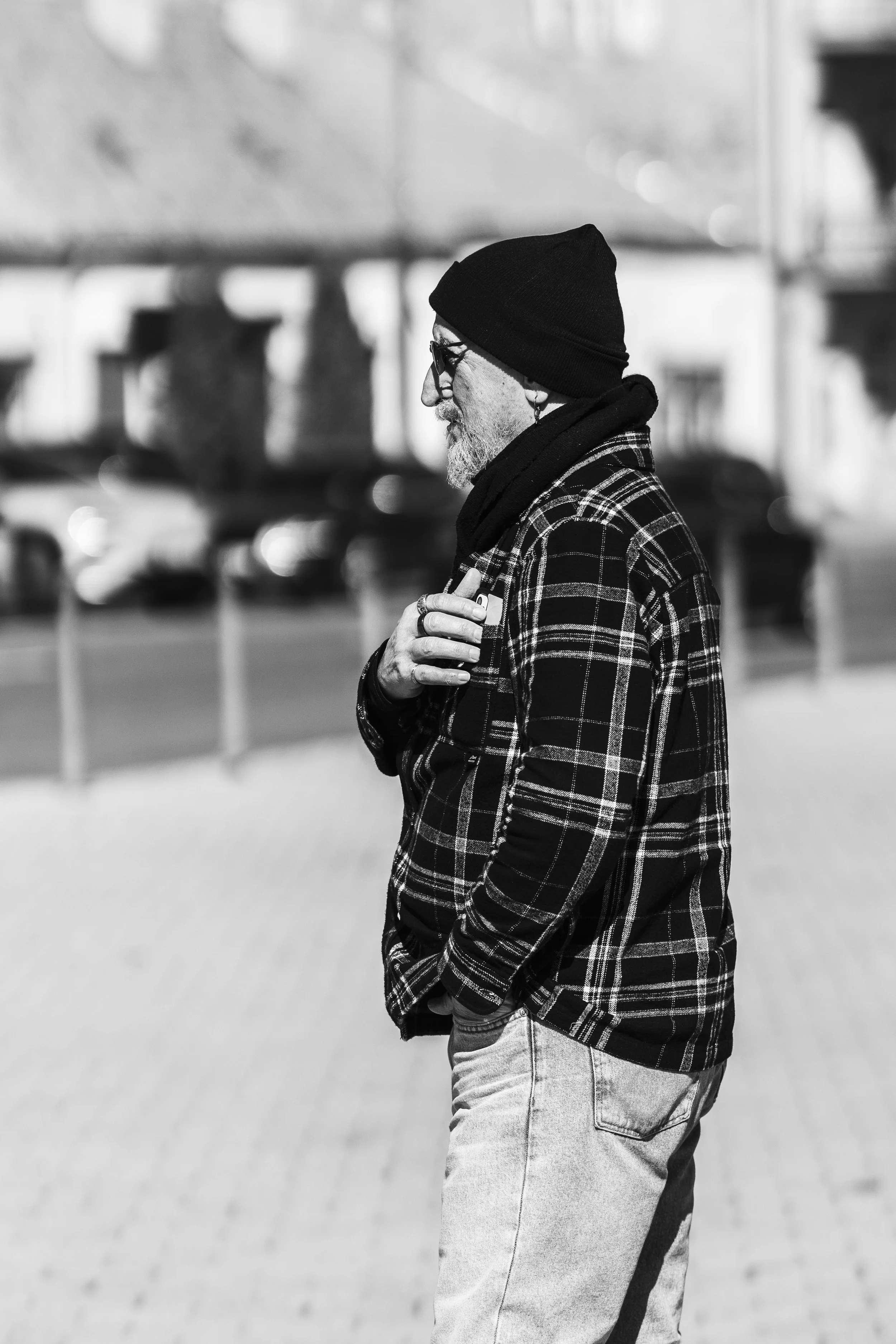 A black and white photo of a man standing outdoors, wearing a black beanie, glasses, a checkered jacket, and a scarf. He is holding a cigarette in his right hand and has his left hand in his pocket, with a blurred background of parked cars and buildi