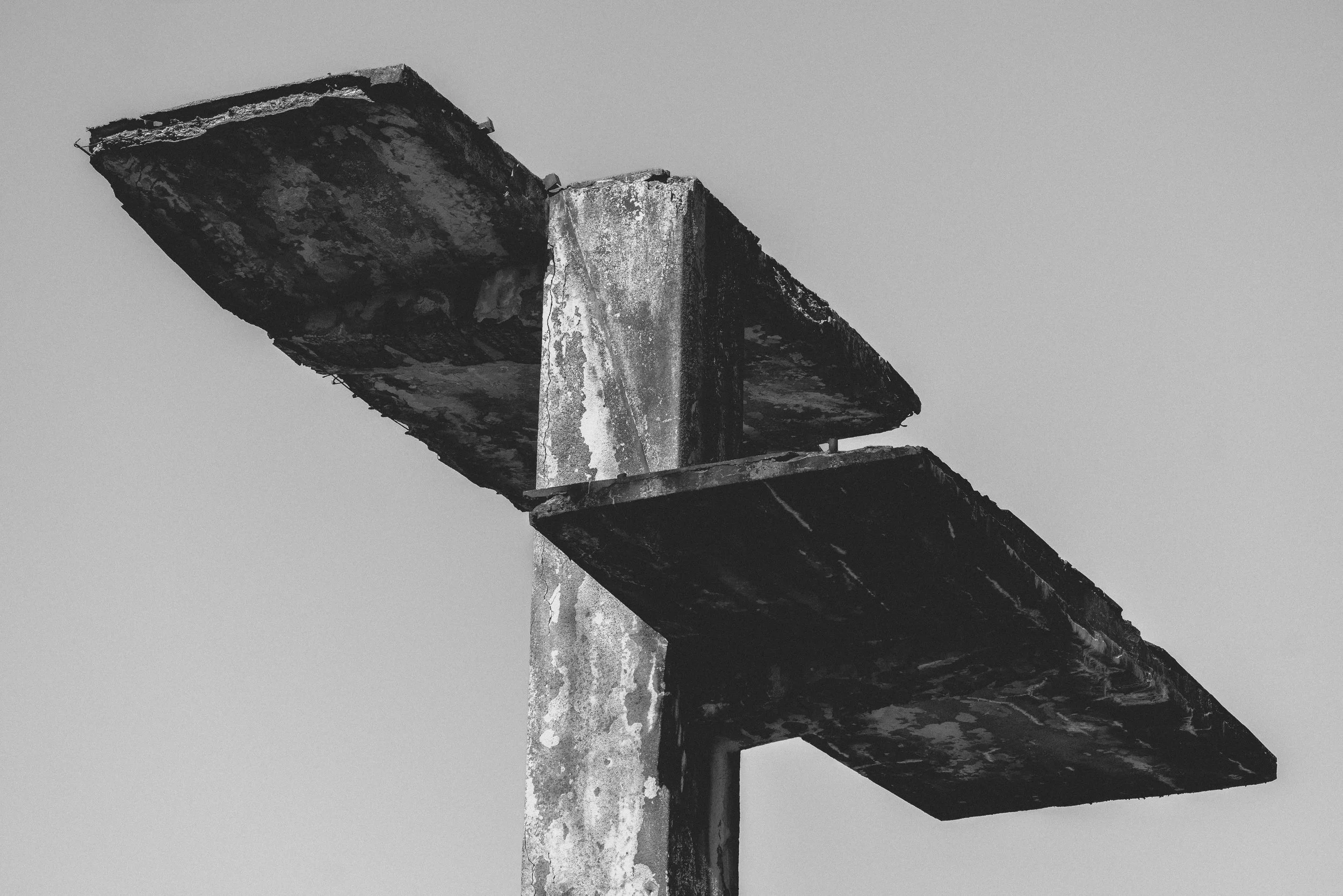 A black and white photo of a weathered, rusted metal structure with two horizontal platforms attached to a vertical pole, against a plain sky background.