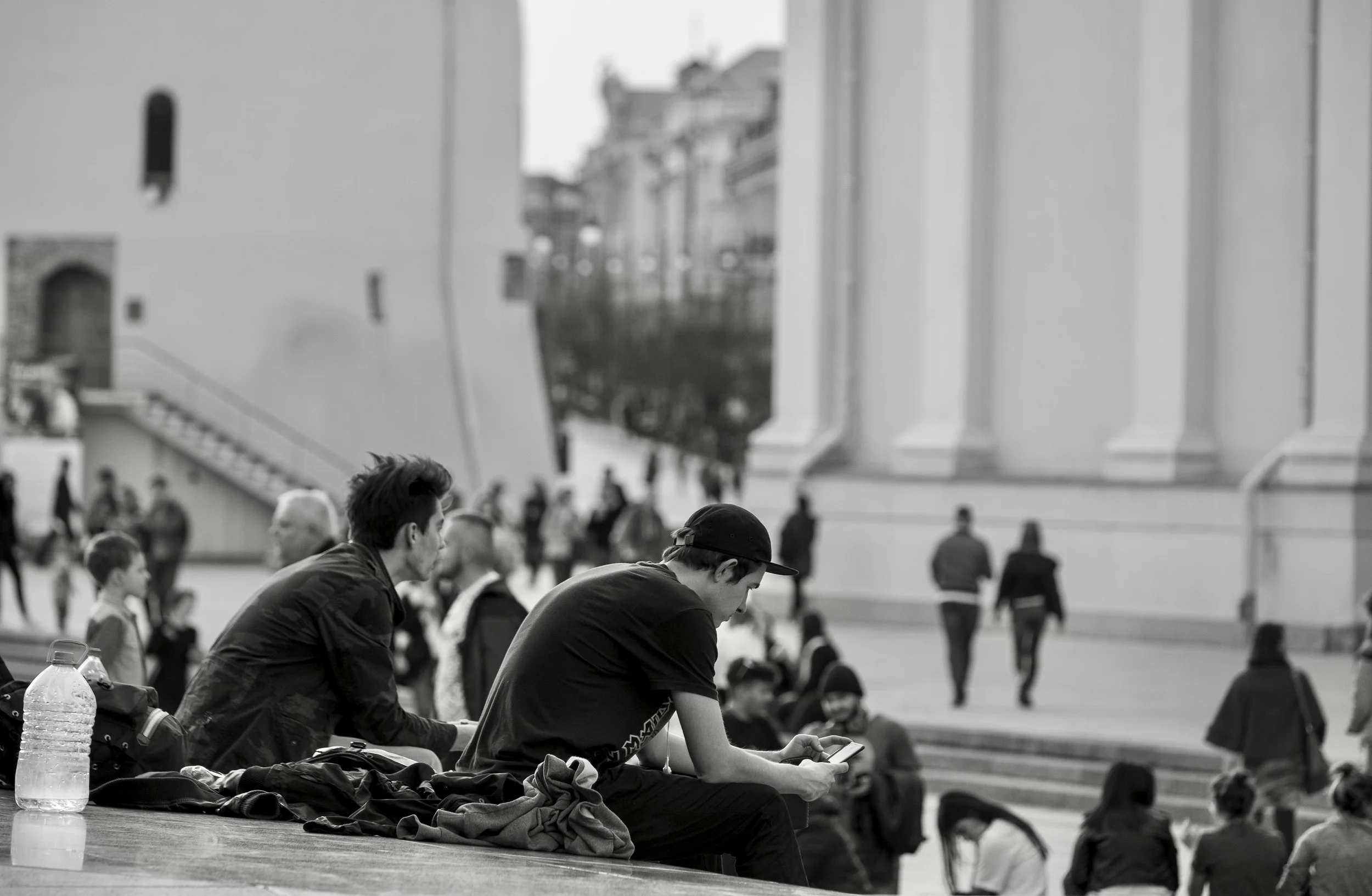 People sitting and walking in an outdoor urban area, with a large building in the background, in black and white.