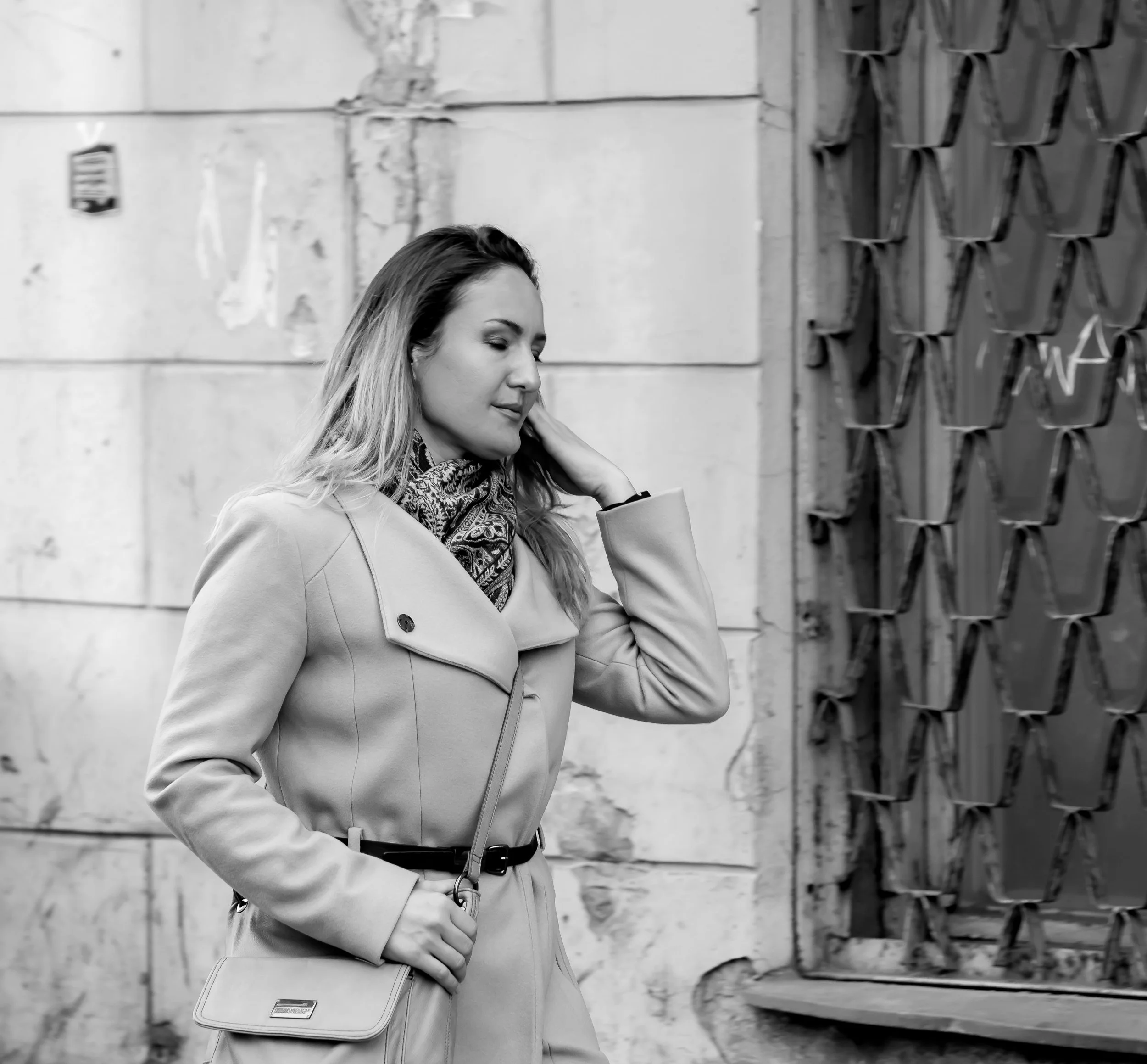 A woman with long hair wearing a trench coat and patterned scarf, walking outdoors and touching her neck, carrying a shoulder bag, in front of a stone wall and decorative metal gate.