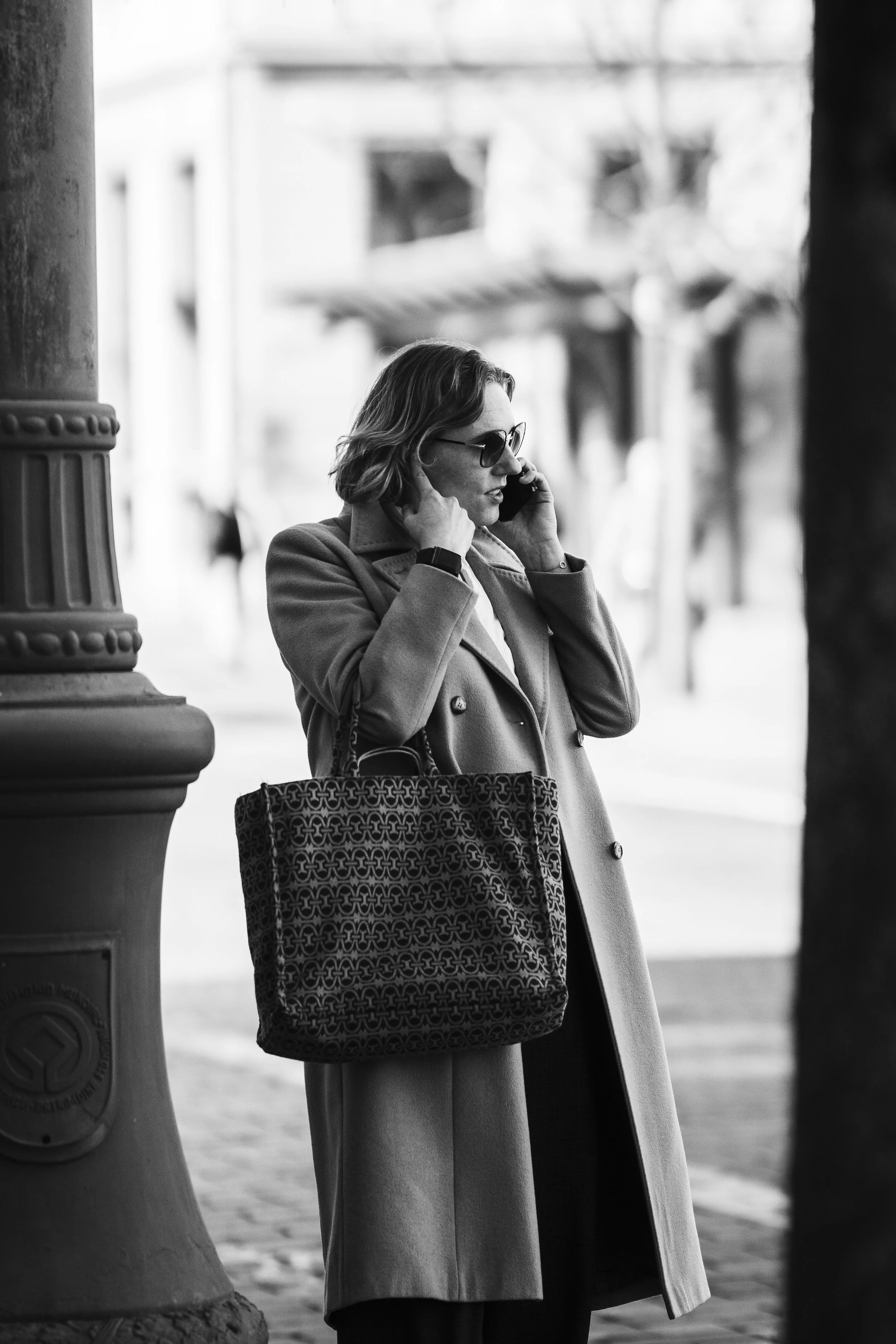 A woman in a trench coat and sunglasses is talking on a cellphone while standing outdoors near a decorative pillar.