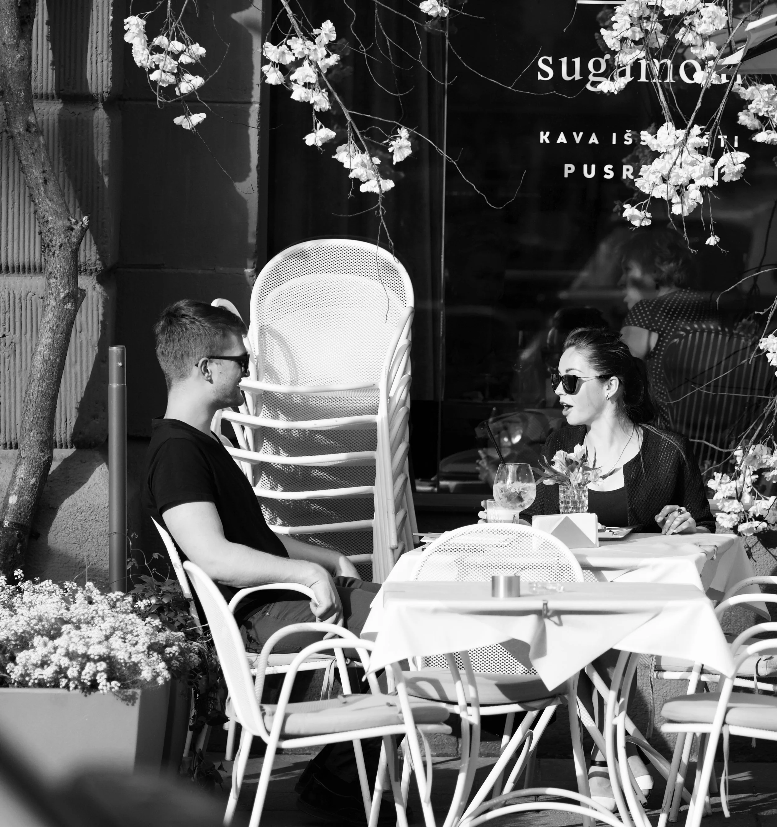 A man and woman are sitting at an outdoor cafe table, engaged in conversation, with the woman wearing sunglasses and drinking a beverage through a straw, and the man also wearing sunglasses, with chairs stacked behind him, and flowers and a tree near