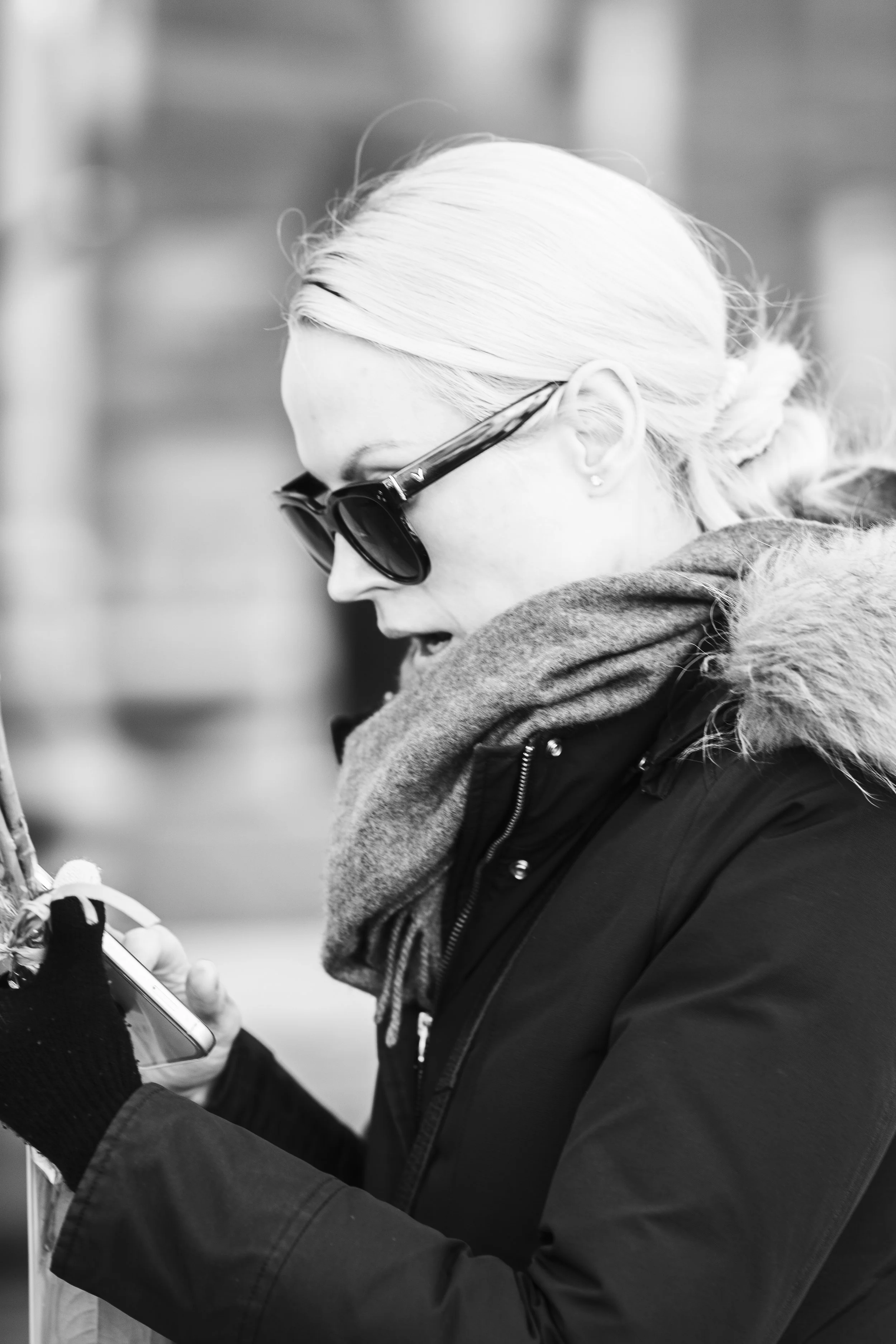 A woman with platinum blonde hair wearing sunglasses, a scarf, and a winter coat looking at her phone.