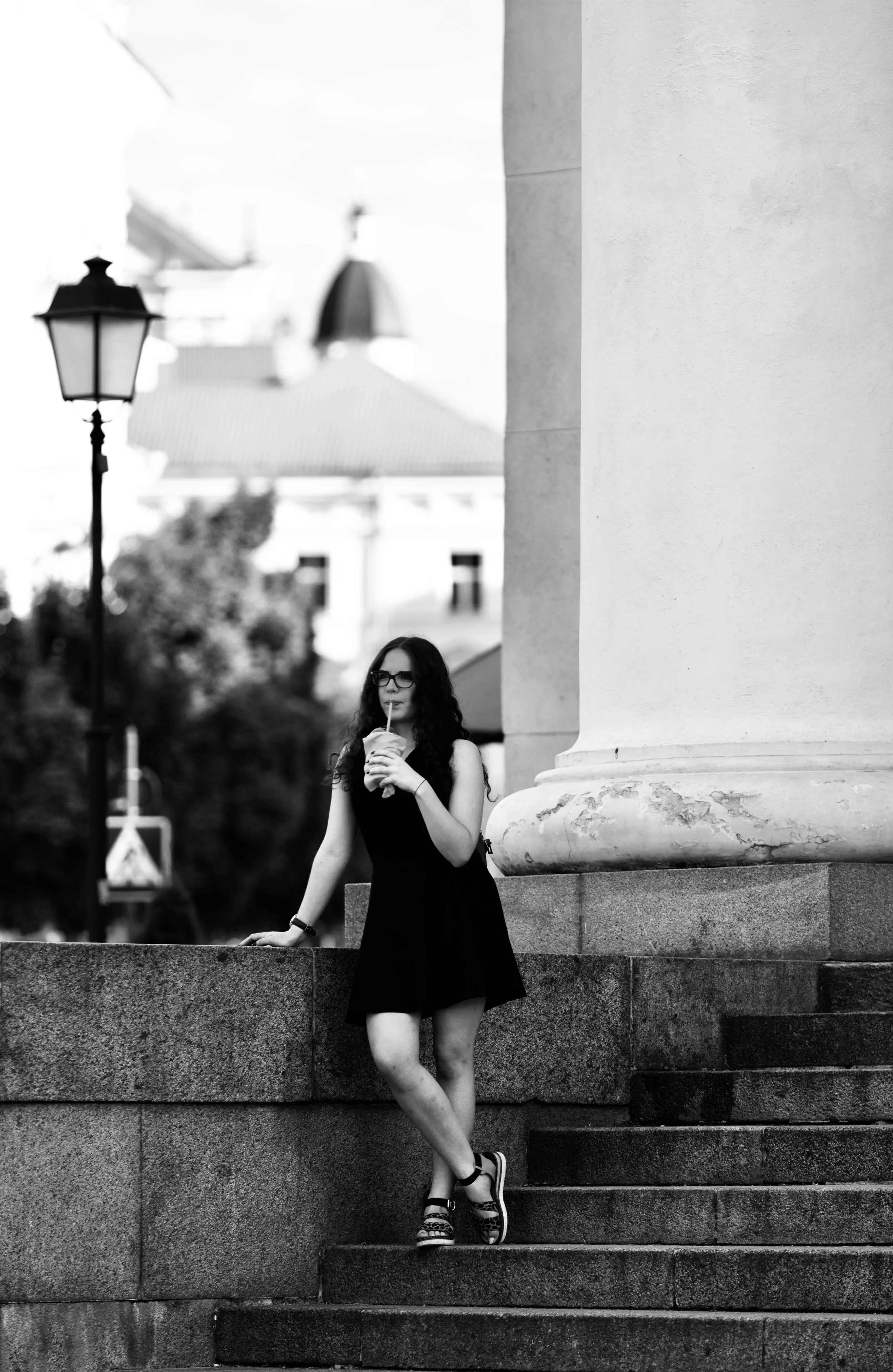 A woman in a black dress and sandals, wearing glasses, is standing on stone steps next to a large column, drinking from a takeaway cup with a straw, in an urban outdoor setting.