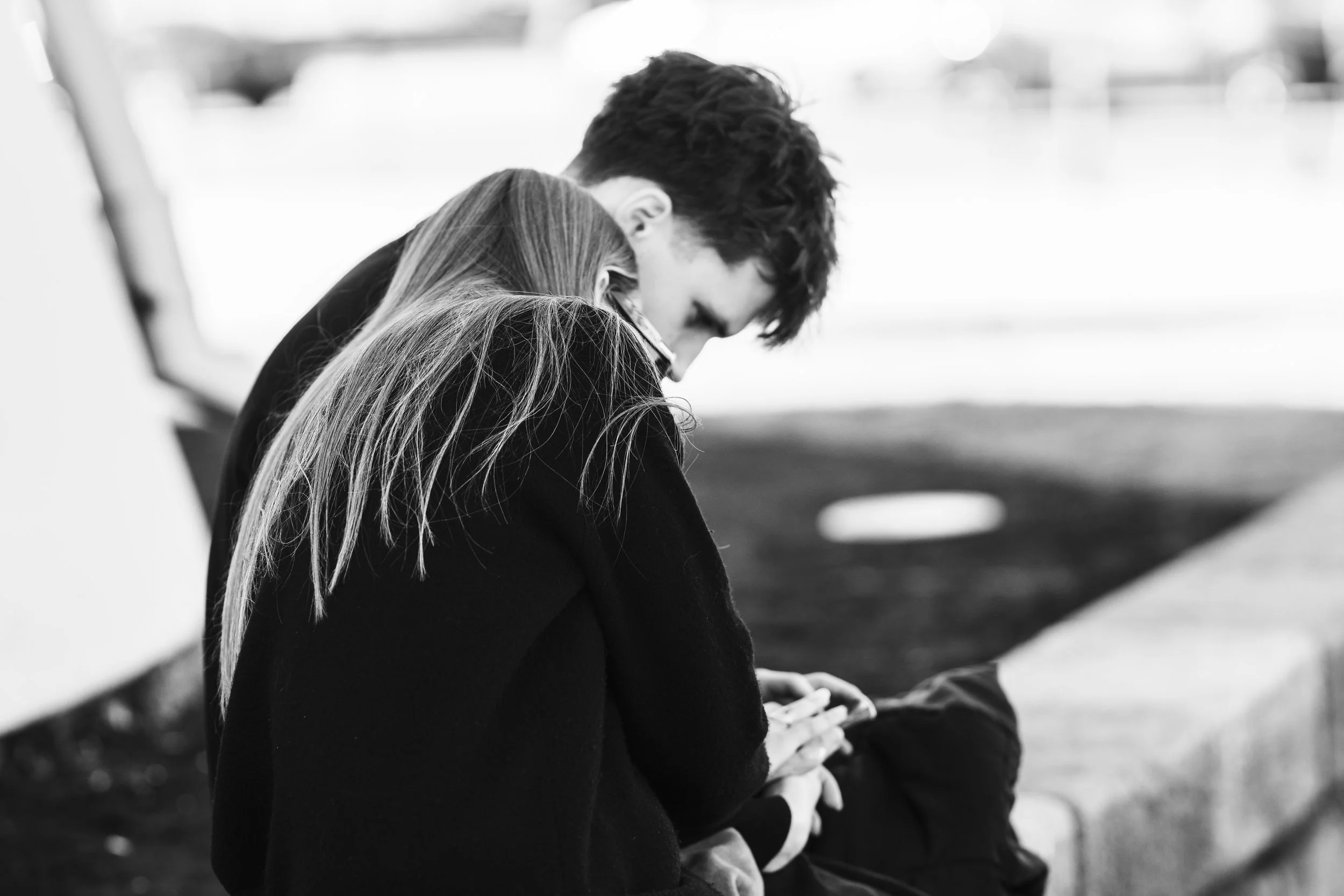 A black and white photo of two young people sitting closely together, looking at a mobile phone. The person with long hair is facing away, while the person with short curly hair is leaning in. They are outdoors, with blurred background elements.