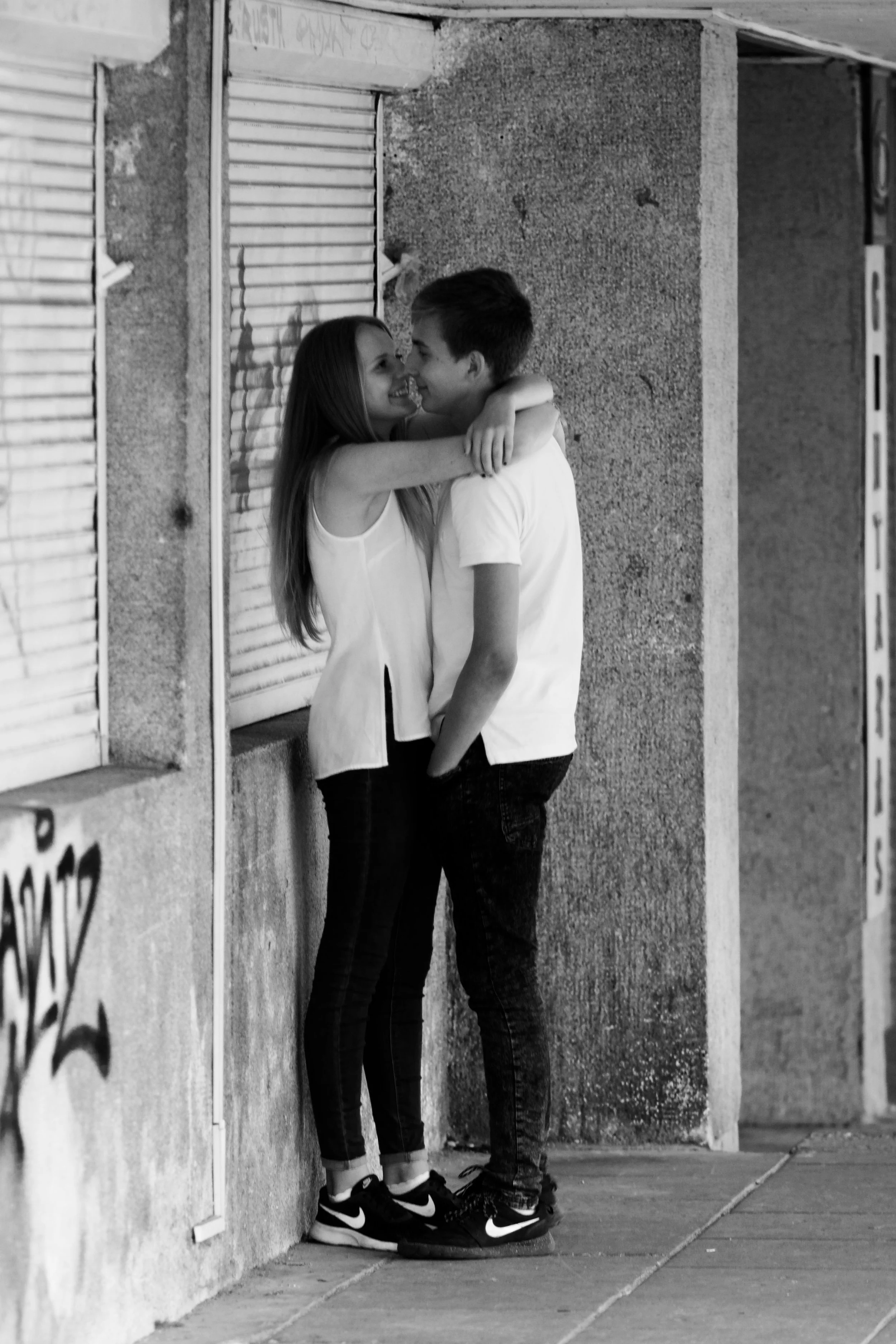 A young couple stands close together at night against a textured wall with graffiti, sharing a happy moment with their foreheads touching and smiling.