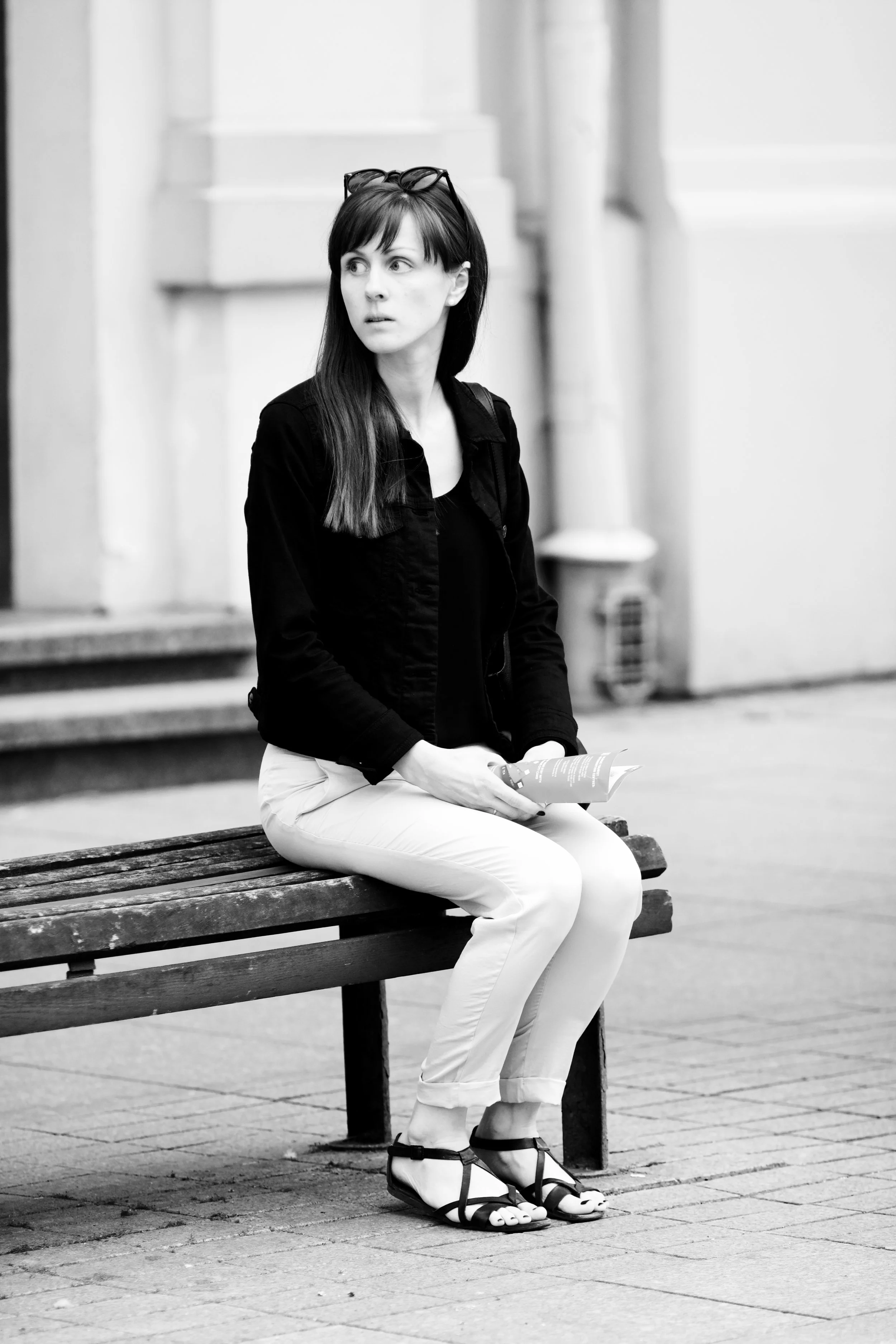 A woman with long dark hair, wearing a black jacket, white pants, and sandals, sitting on a wooden bench while holding a pamphlet or brochure in an urban setting.