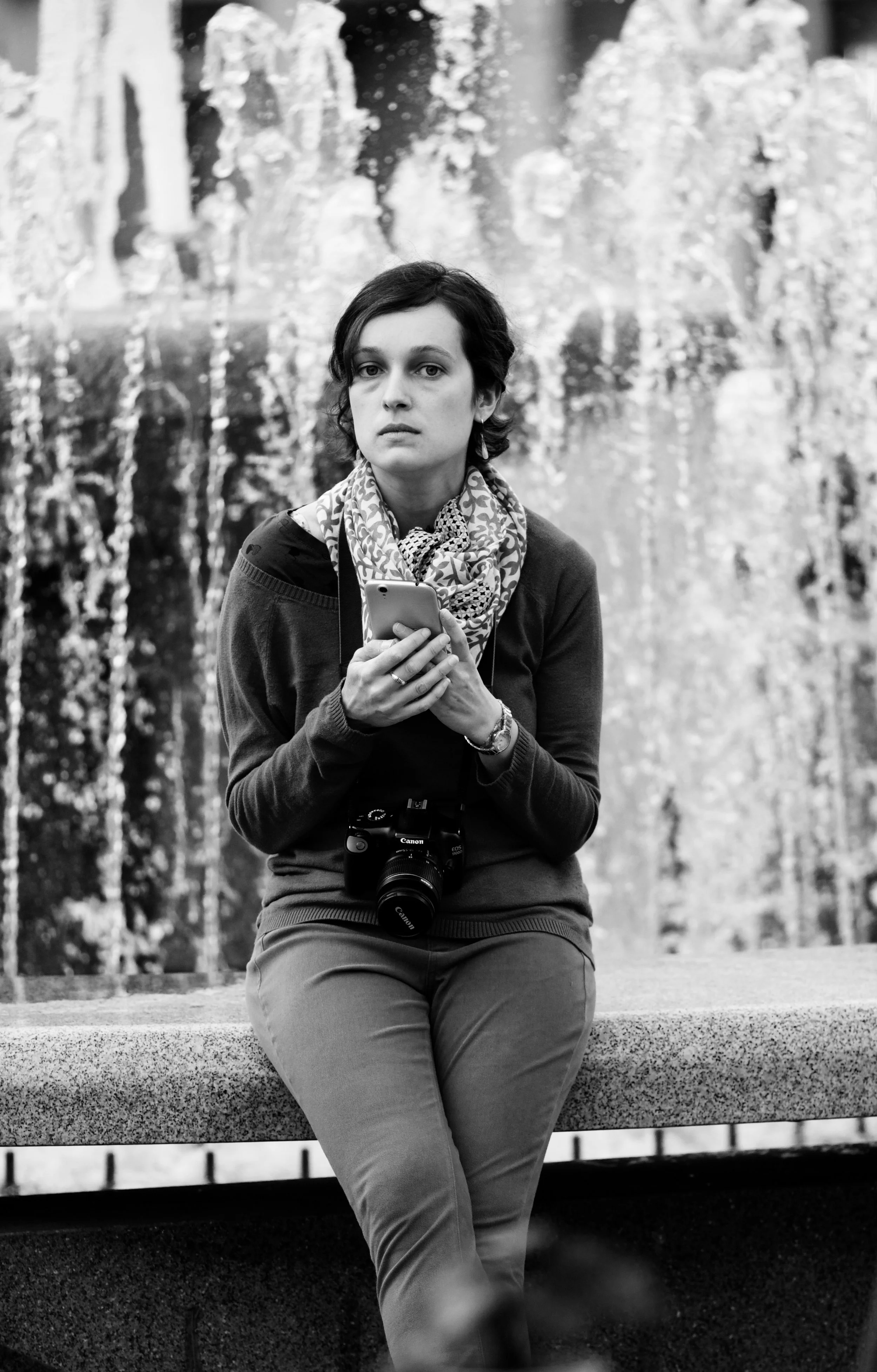 A woman with short dark hair, wearing a patterned scarf and a dark sweater, is sitting on a bench holding a smartphone. She has a camera hanging around her neck and is standing in front of a fountain.