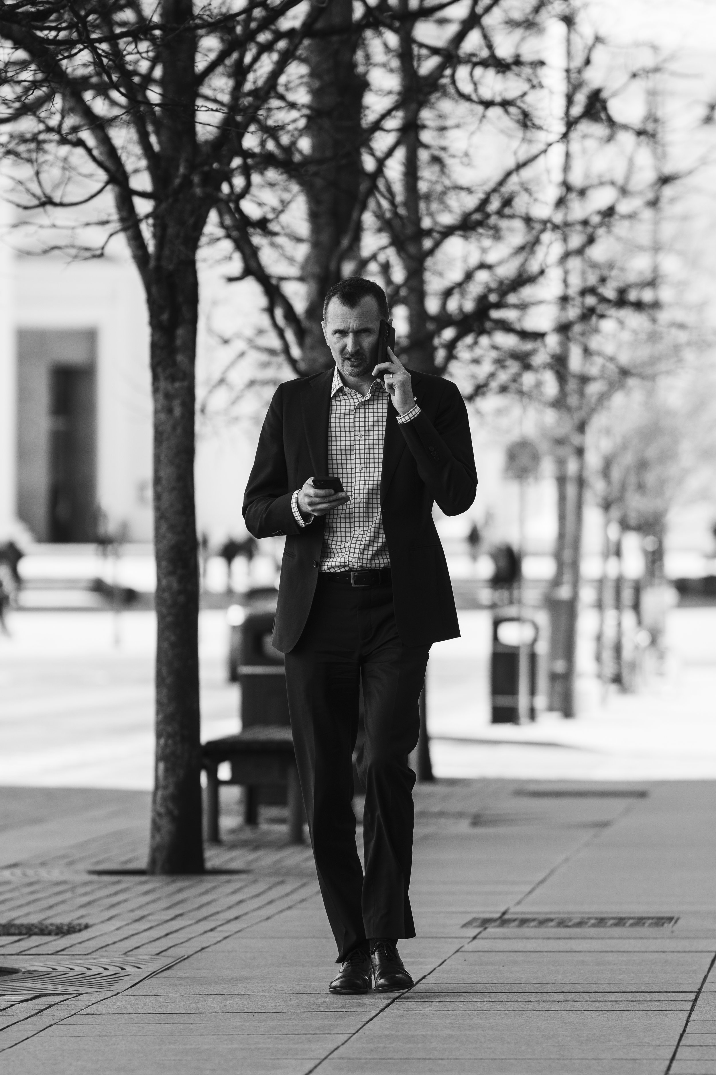 A man in business attire walking on a city sidewalk while talking on his cell phone, with trees and benches in the background.