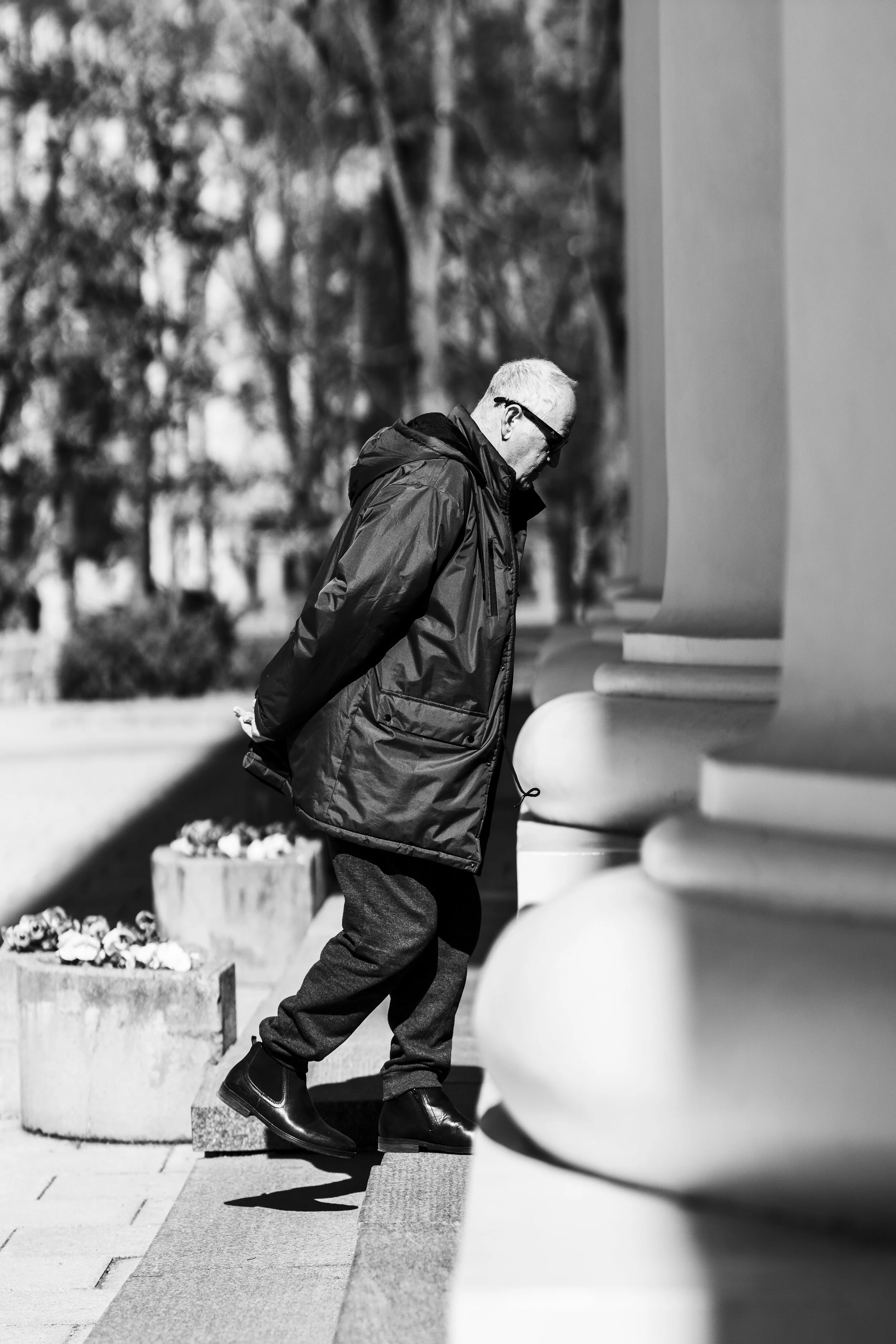 An older man with gray hair, wearing sunglasses, a dark jacket, and dark trousers, walking up the steps of a building with large columns on a sunny day.
