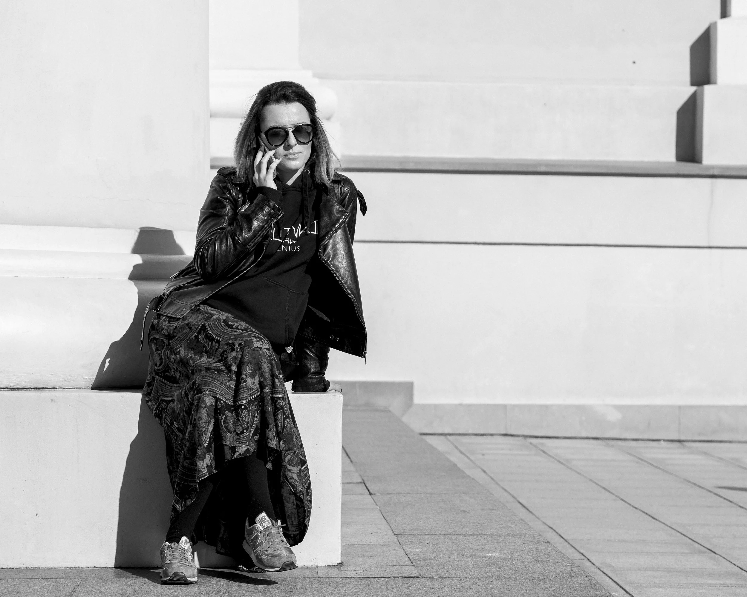 A young woman with sunglasses, leather jacket, and patterned skirt sitting on a ledge, talking on a cellphone, outdoors with a minimalist architectural background.