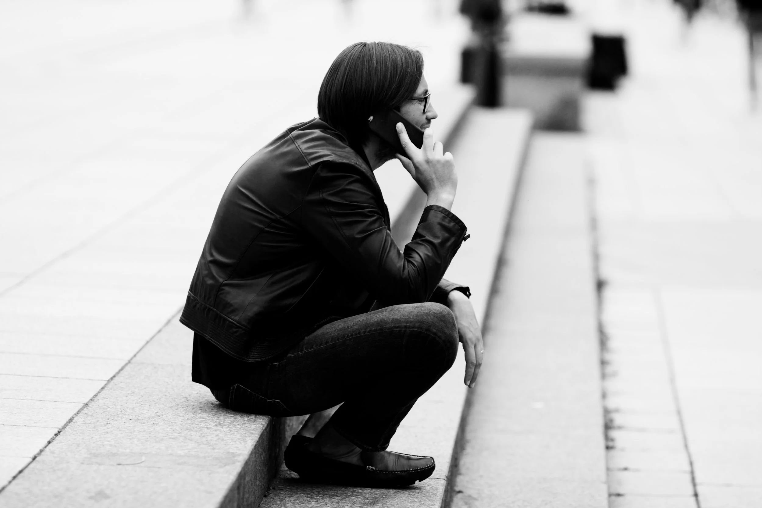 A woman with glasses and a leather jacket sitting on a bench, squatting, and talking on her cell phone.