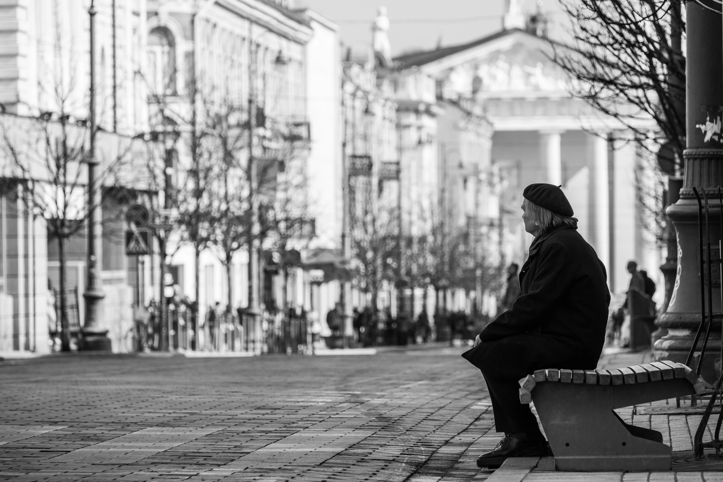A black-and-white photo of an elderly woman sitting alone on a park bench in an urban setting with buildings and trees in the background.