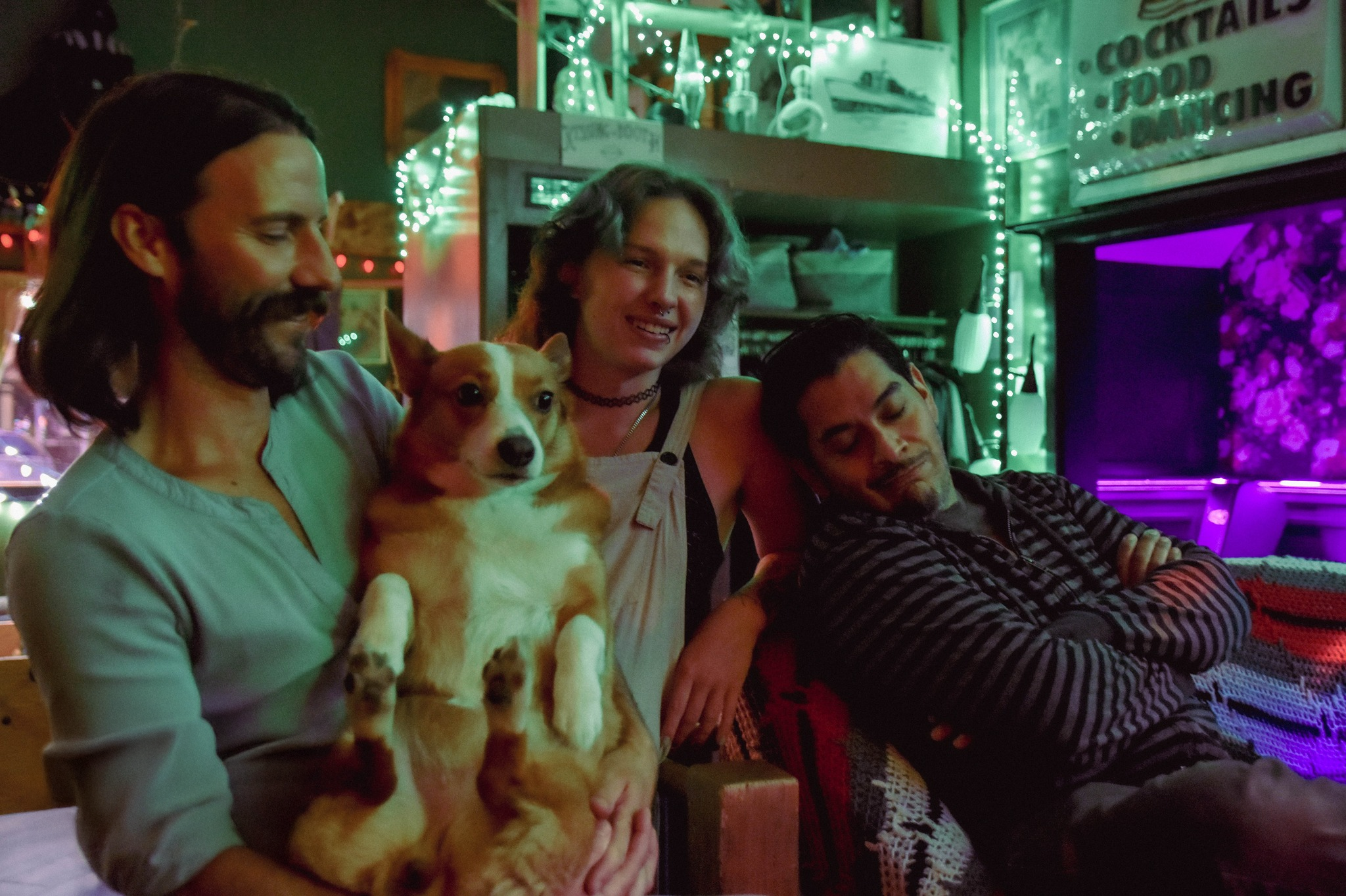 Three people and a dog sitting together in a cozy, colorful, neon-lit room. The man on the left has long dark hair and a beard, holding a small dog. The woman in the middle has shoulder-length hair and is smiling. The man on the right has short dark hair and is resting with his eyes closed. The room has green and purple neon lighting, string lights, and signs, creating a relaxed, festive atmosphere.