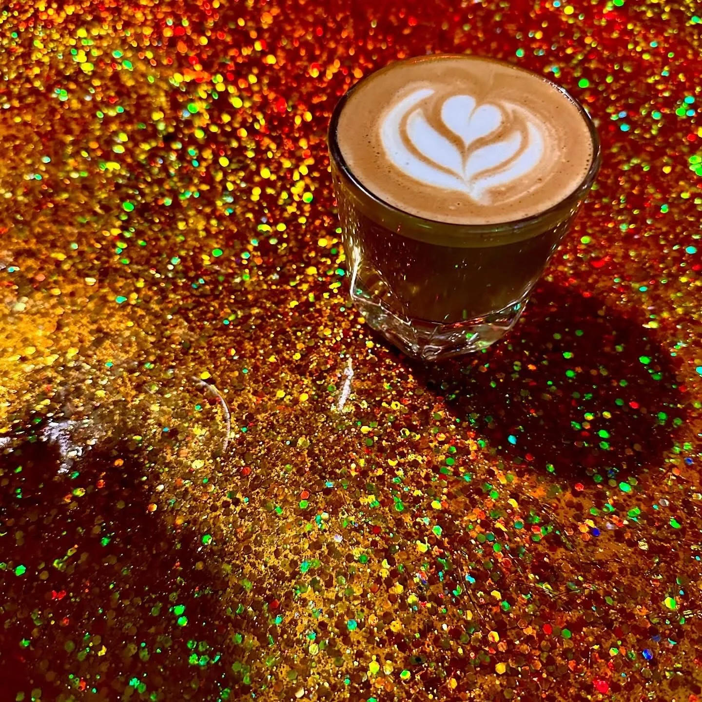 A glass of coffee or latte with latte art in the shape of a heart on top, placed on a shimmering, colorful, glittery surface.