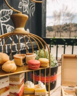 A two-tiered gold dessert stand with assorted macarons, croissants, and cake slices on a table outside, with a black decorative wall and a balcony in the background.